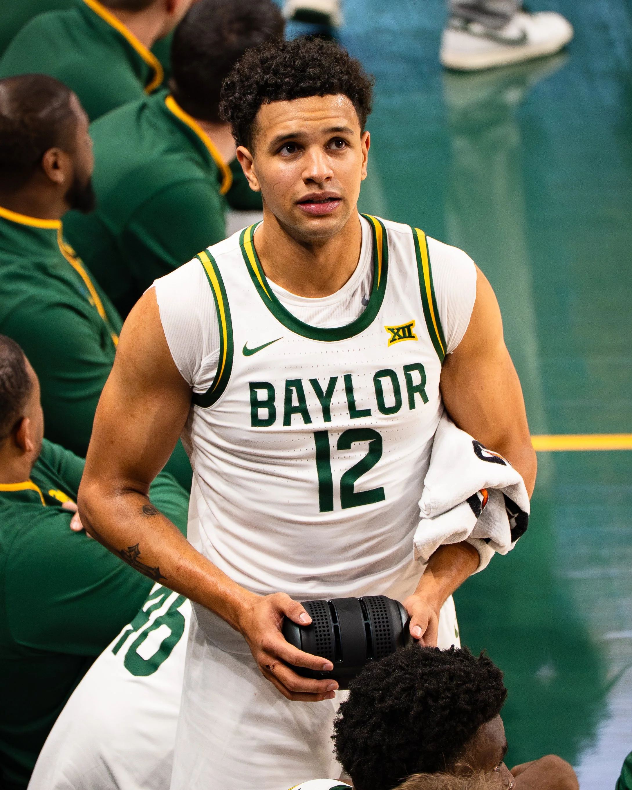 A basketball player in a white Baylor uniform holds a microphone while sitting on the bench during a game, surrounded by teammates and staff.