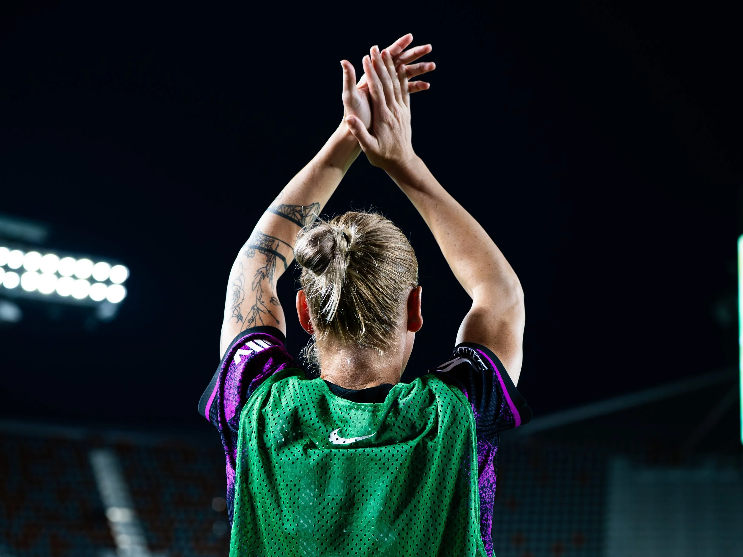 Person in a green sports jersey with purple and black accents raising their hands during a sports event, dark background with stadium lights.
