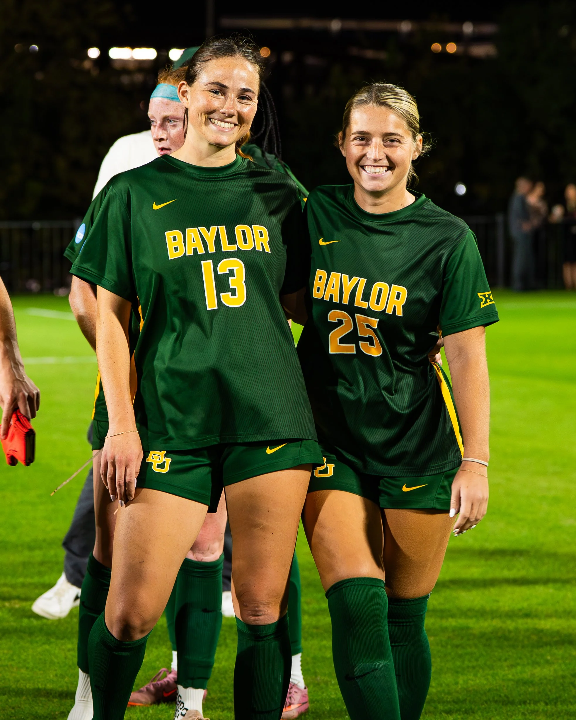 Two female soccer players in green jerseys standing on a field, smiling, with a third person in the background. The jerseys say 'BAYLOR' and have yellow Nike logos.