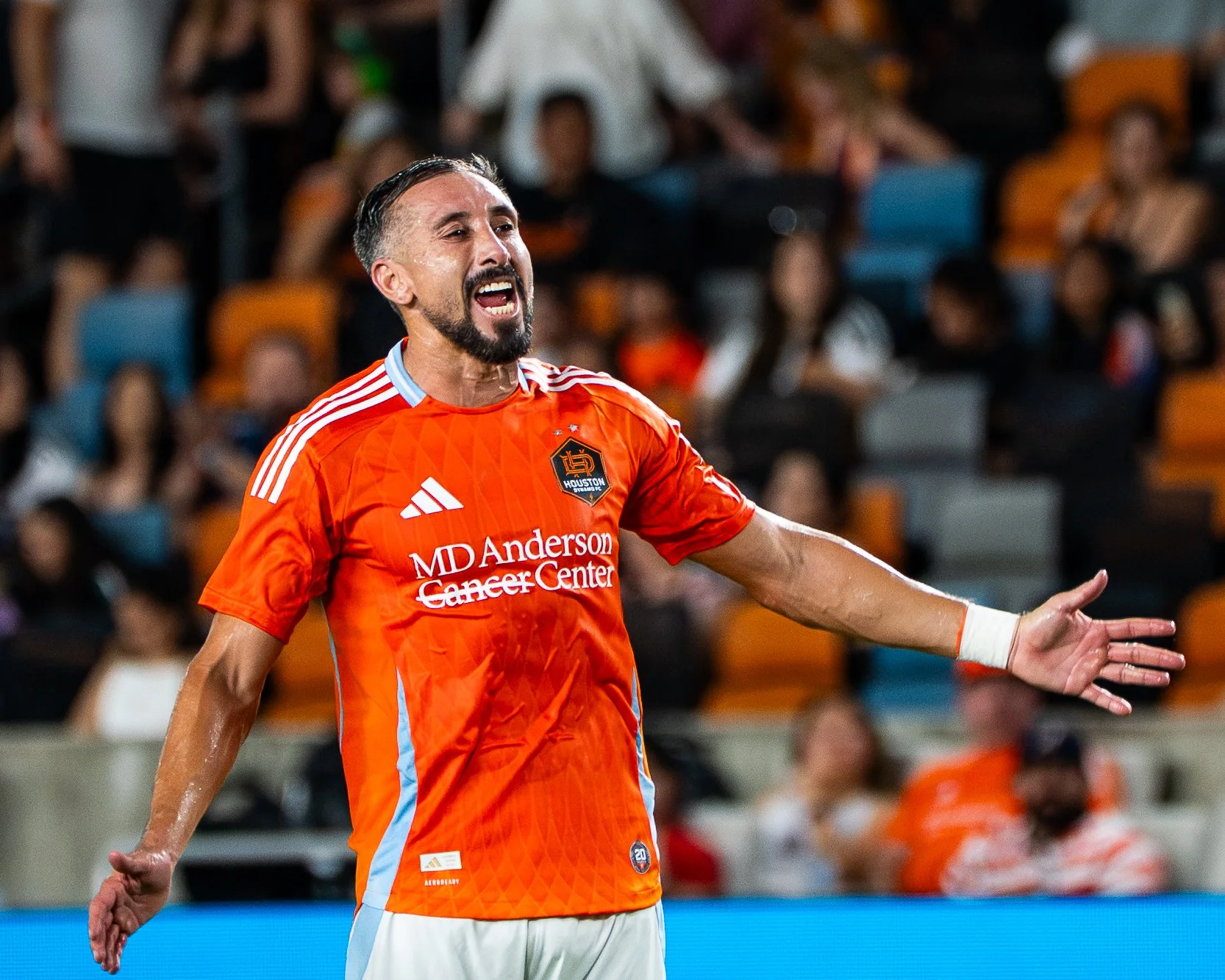A male soccer player with dark hair, beard, and light skin, wearing an orange Houston Dynamo jersey, is celebrating on the field with a background of fans in stadium seats.
