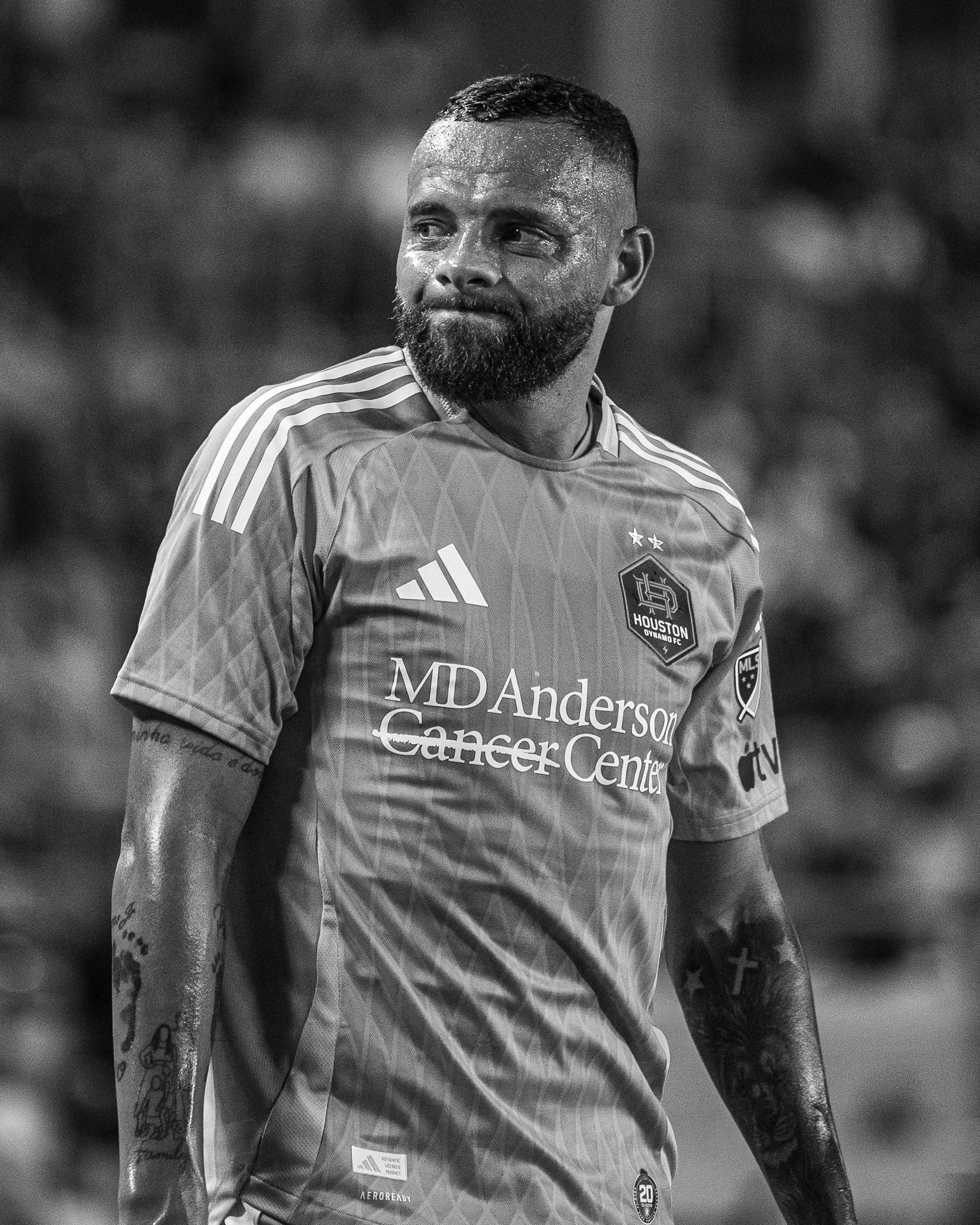 A football player with a beard and short hair, wearing a Houston Dynamo FC jersey with the MDA Anderson Cancer Center logo, sweating and looking to his right.