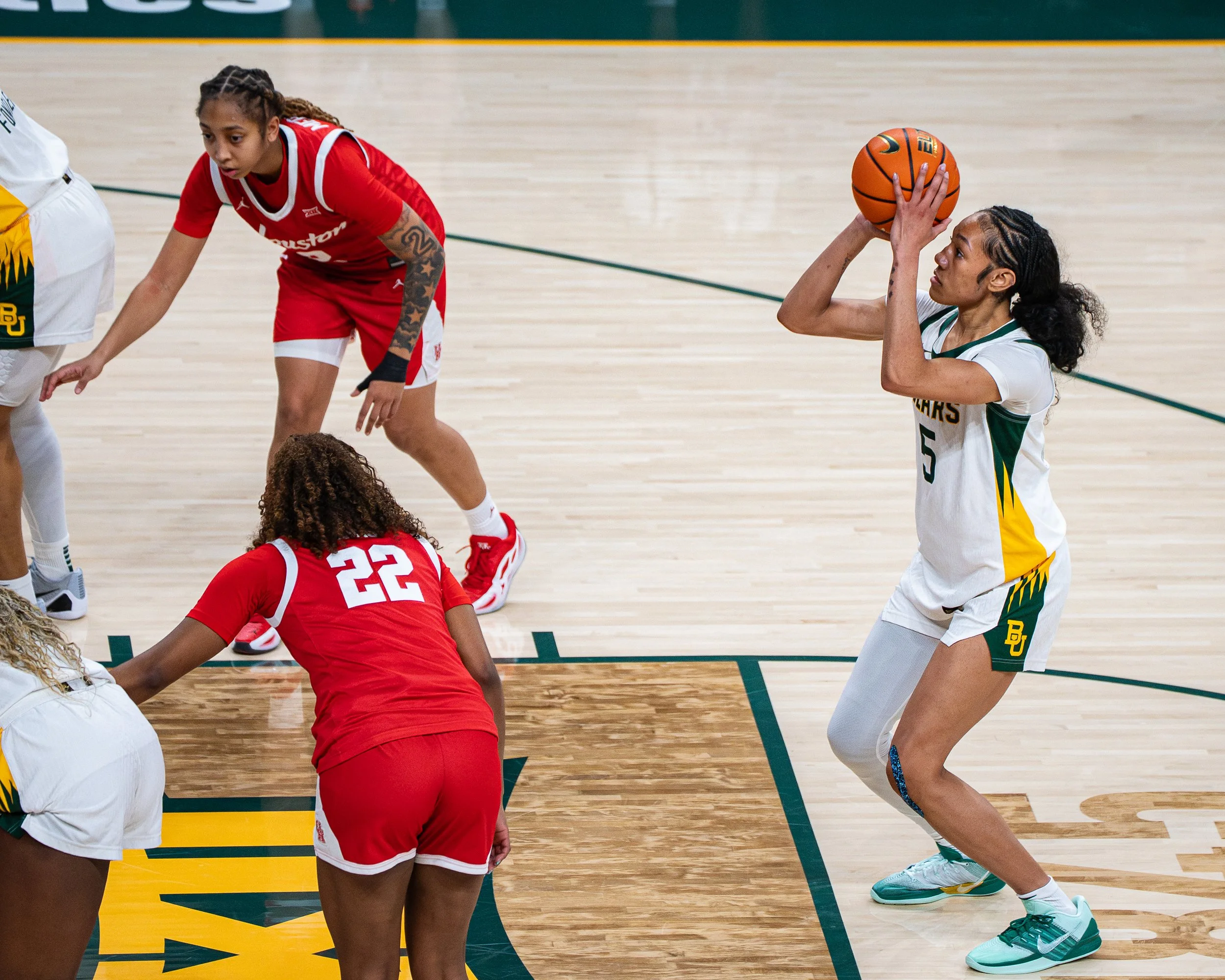 A women's basketball game with players on the court, one player in white about to shoot the ball and another in red on the ground attempting to block.