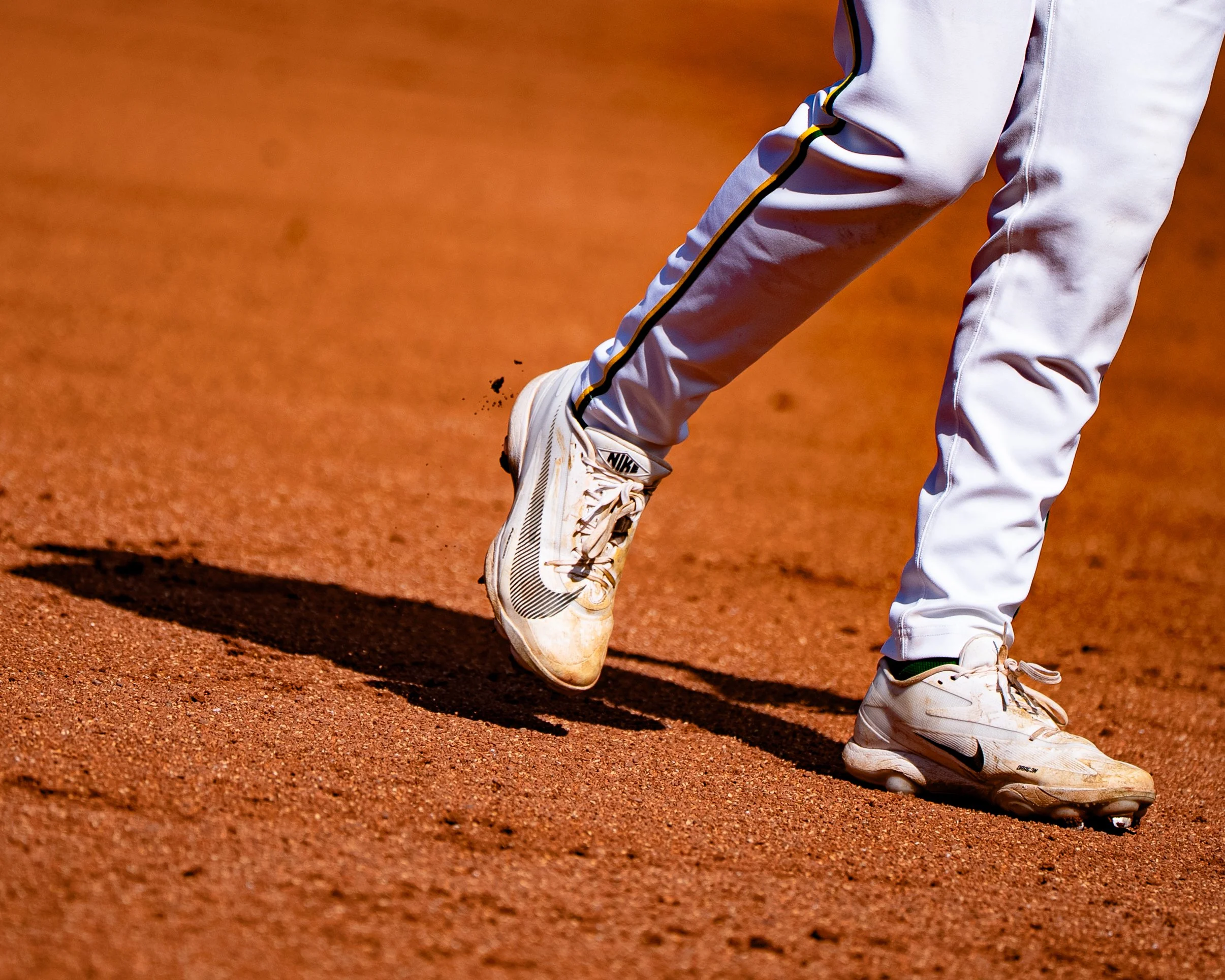 Close-up of a baseball player's feet on a dirt field, wearing white Nike cleats and white pants with yellow and black piping.