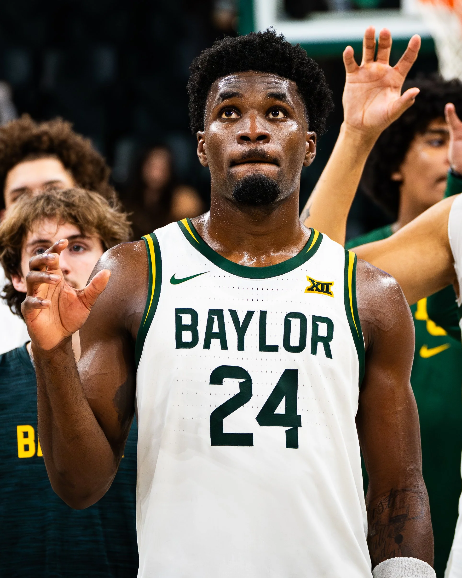 A basketball player wearing a Baylor jersey with the number 24, looking upwards with a focused expression, surrounded by fellow team members.