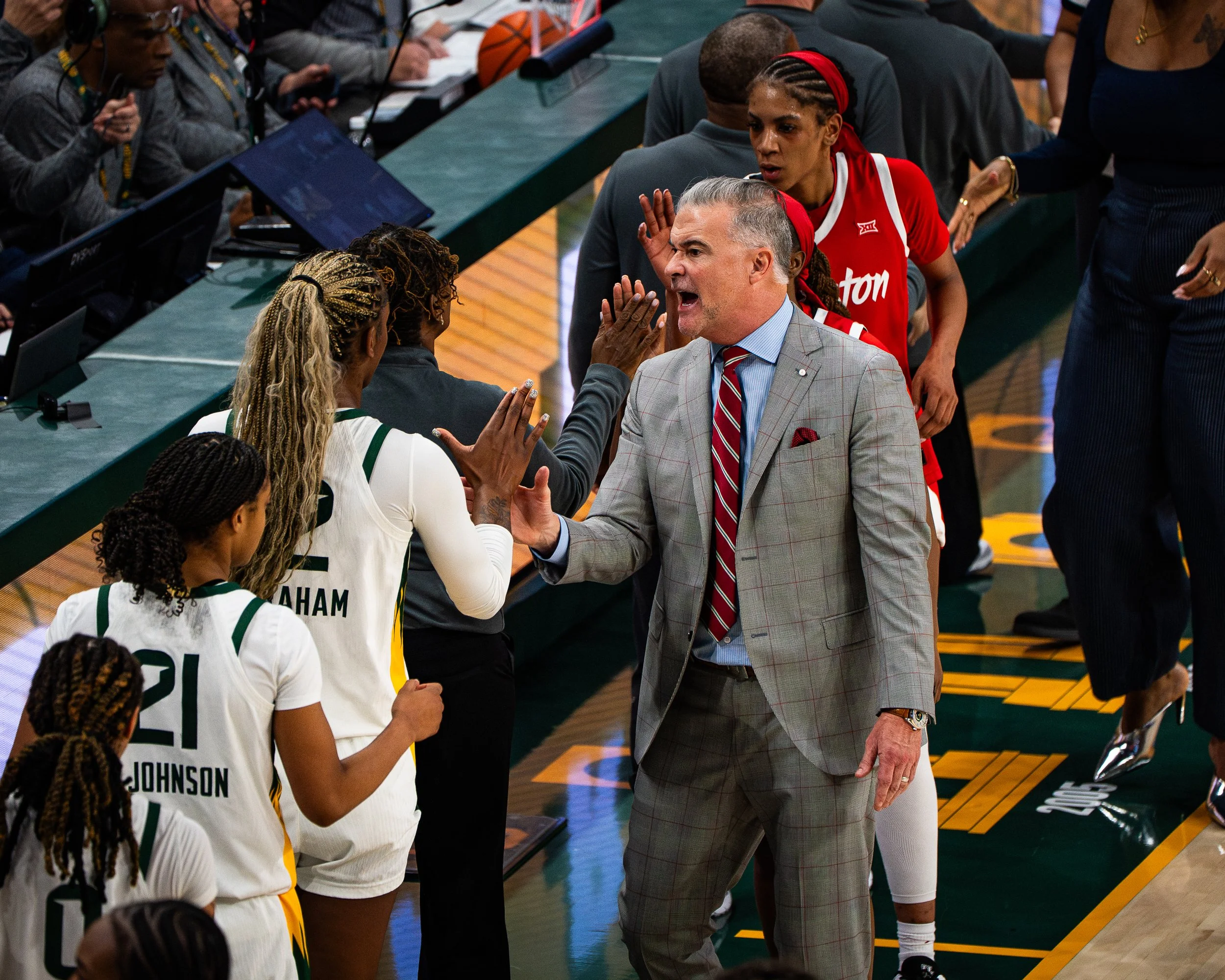 An indoor basketball setting with players and officials. A coach in a suit is speaking passionately to players in white and green uniforms. The players are listening, some with their hands raised, and there are electronics and basketballs on the tabl