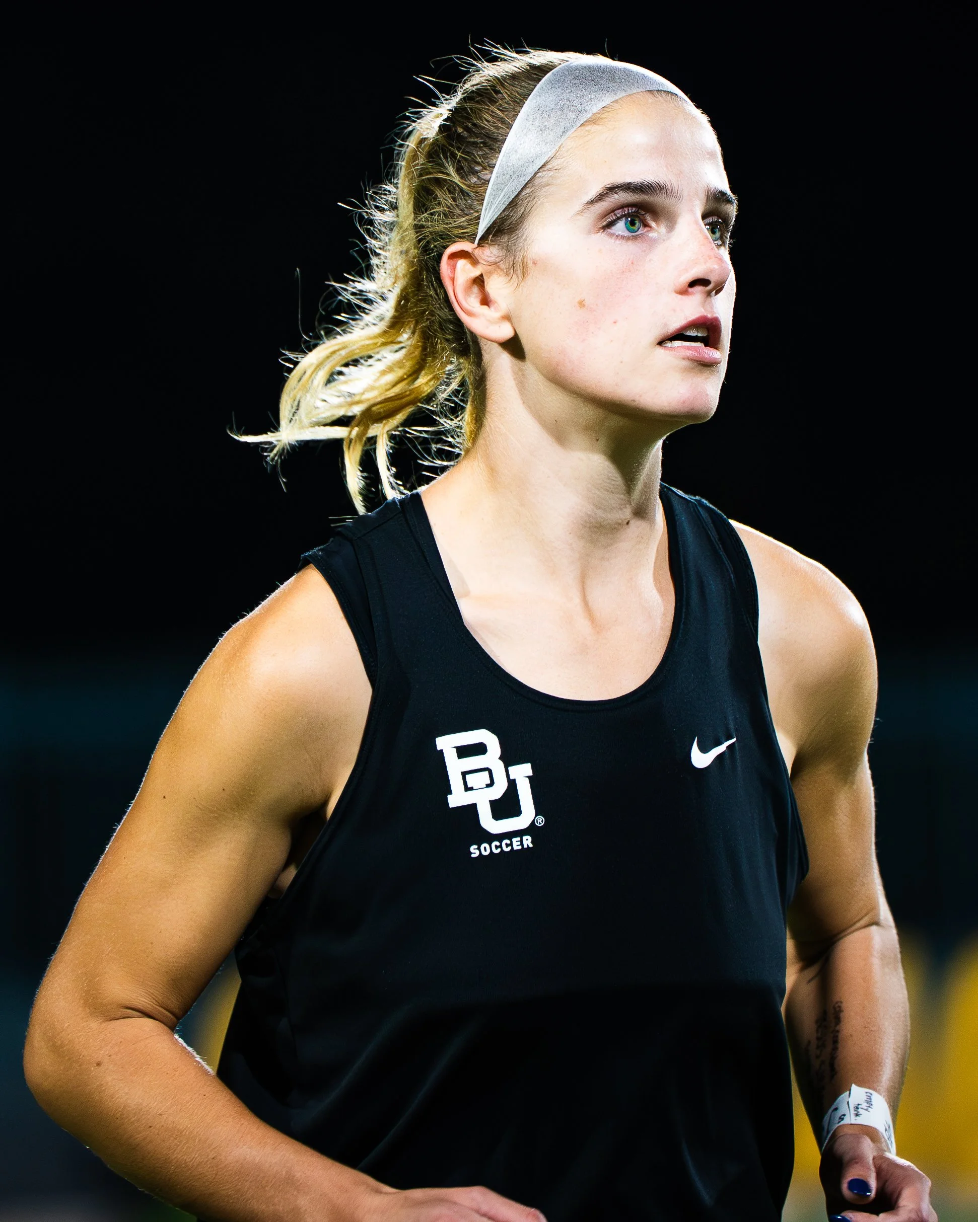 Close-up of a female athlete with blonde hair, wearing a headband and a black sports tank top with a logo, running outdoors at night.