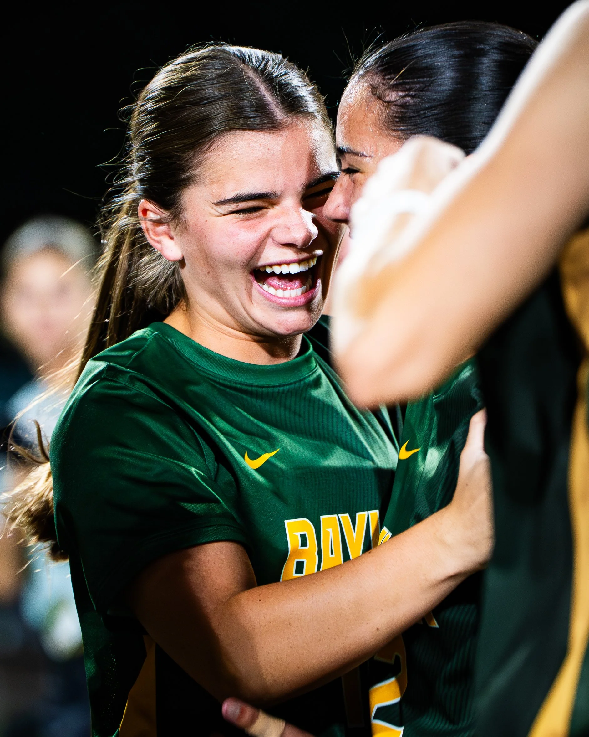 Two female athletes, one in green jersey and the other in black and yellow jersey, are in a close embrace after a sports match, sharing an emotional moment of joy.