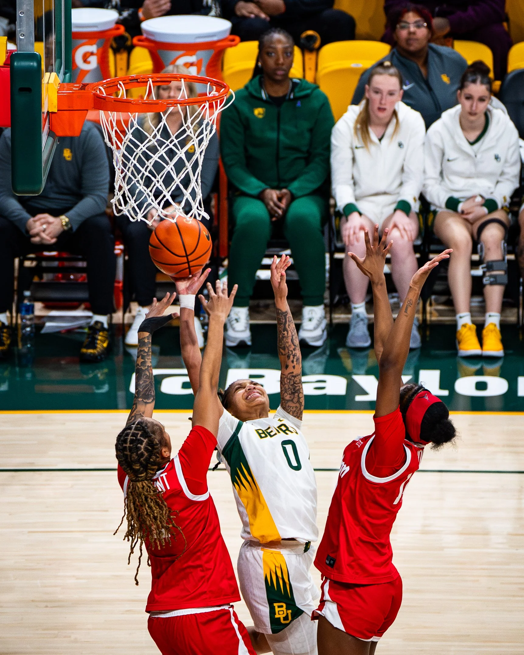 A basketball game in progress with players competing for the ball near the hoop, with players and spectators seated in the background.