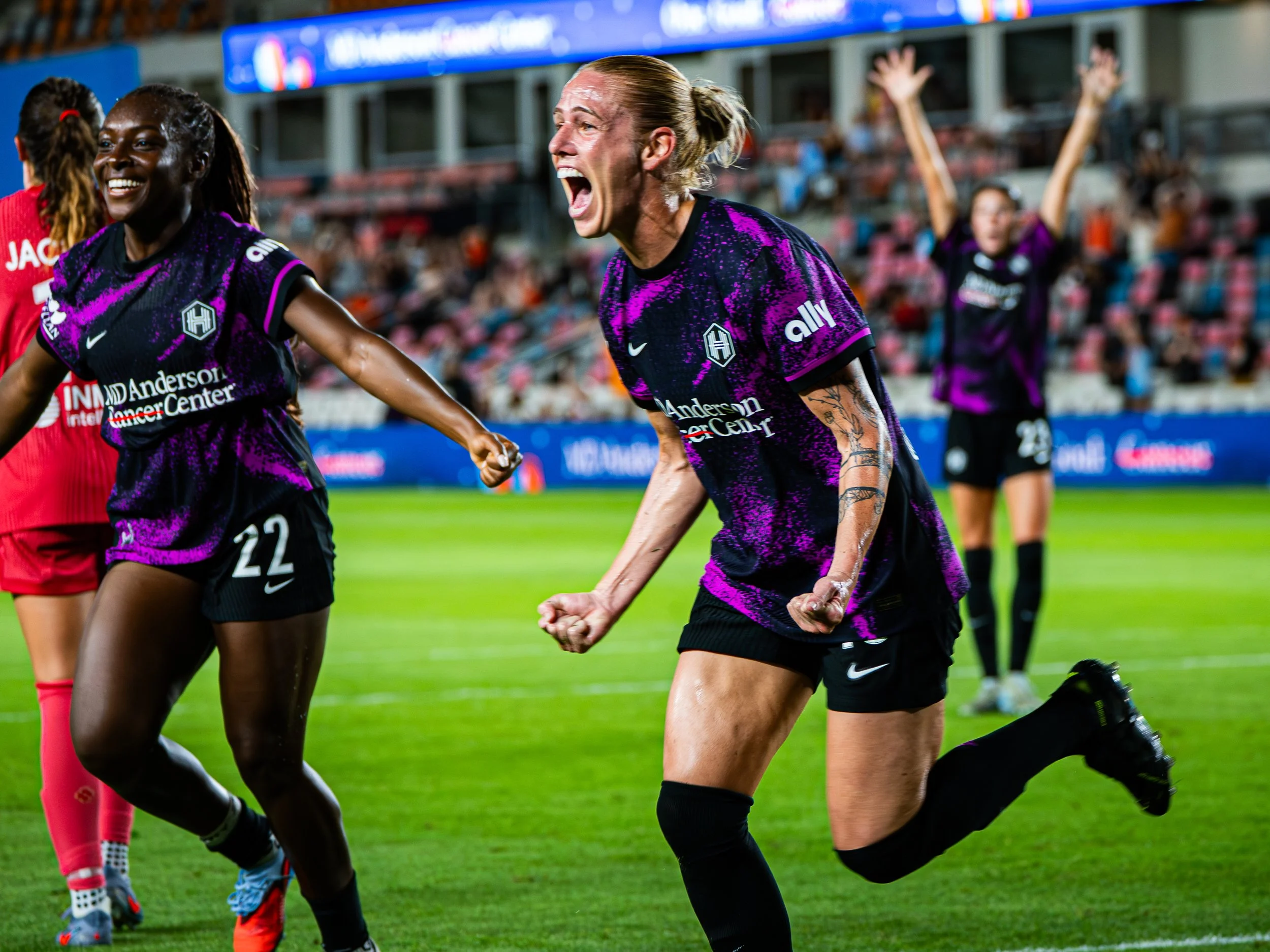 Women celebrating a goal during a soccer match, wearing purple and black uniforms, on a green field with spectators in the background.