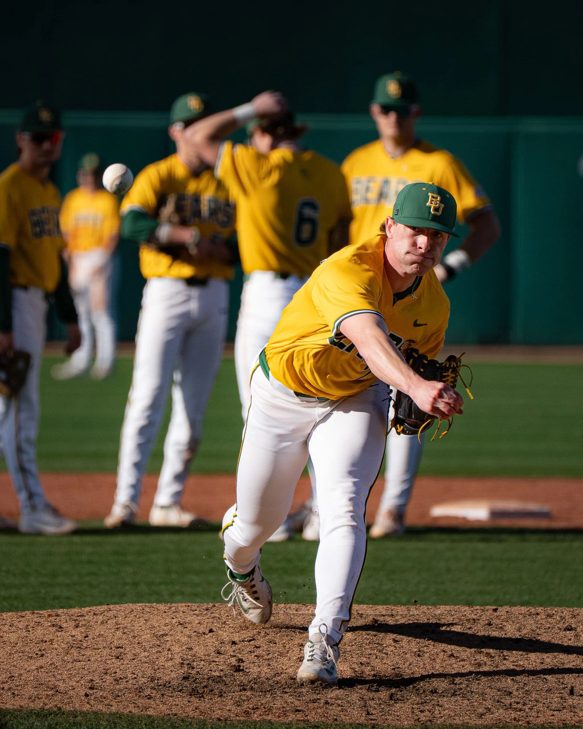 A baseball pitcher in yellow and white uniform on the mound, with teammates in the background during a game.