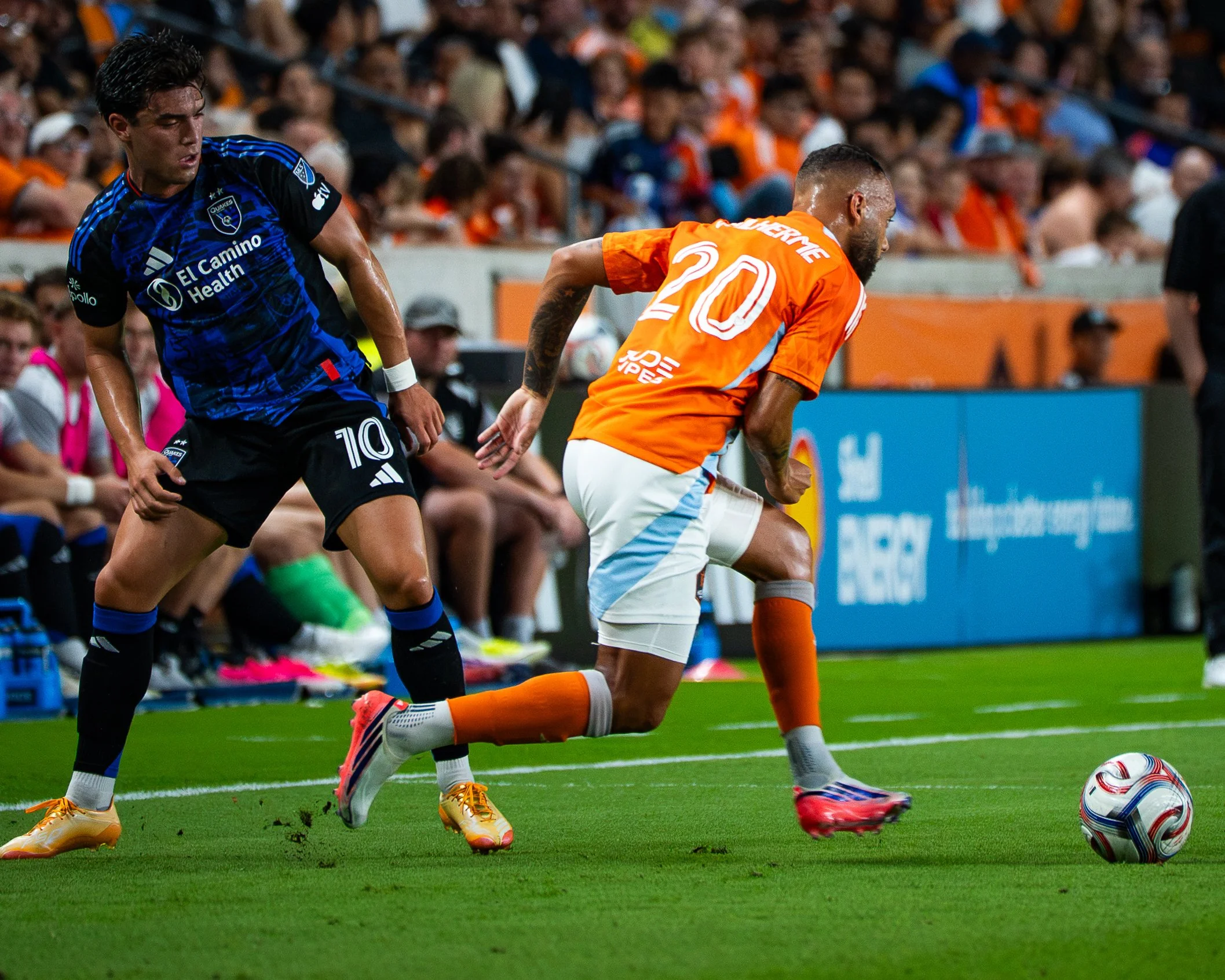 Two soccer players, one wearing a black and blue uniform and the other in an orange uniform, are chasing a soccer ball on a green field with a crowd in the stands.