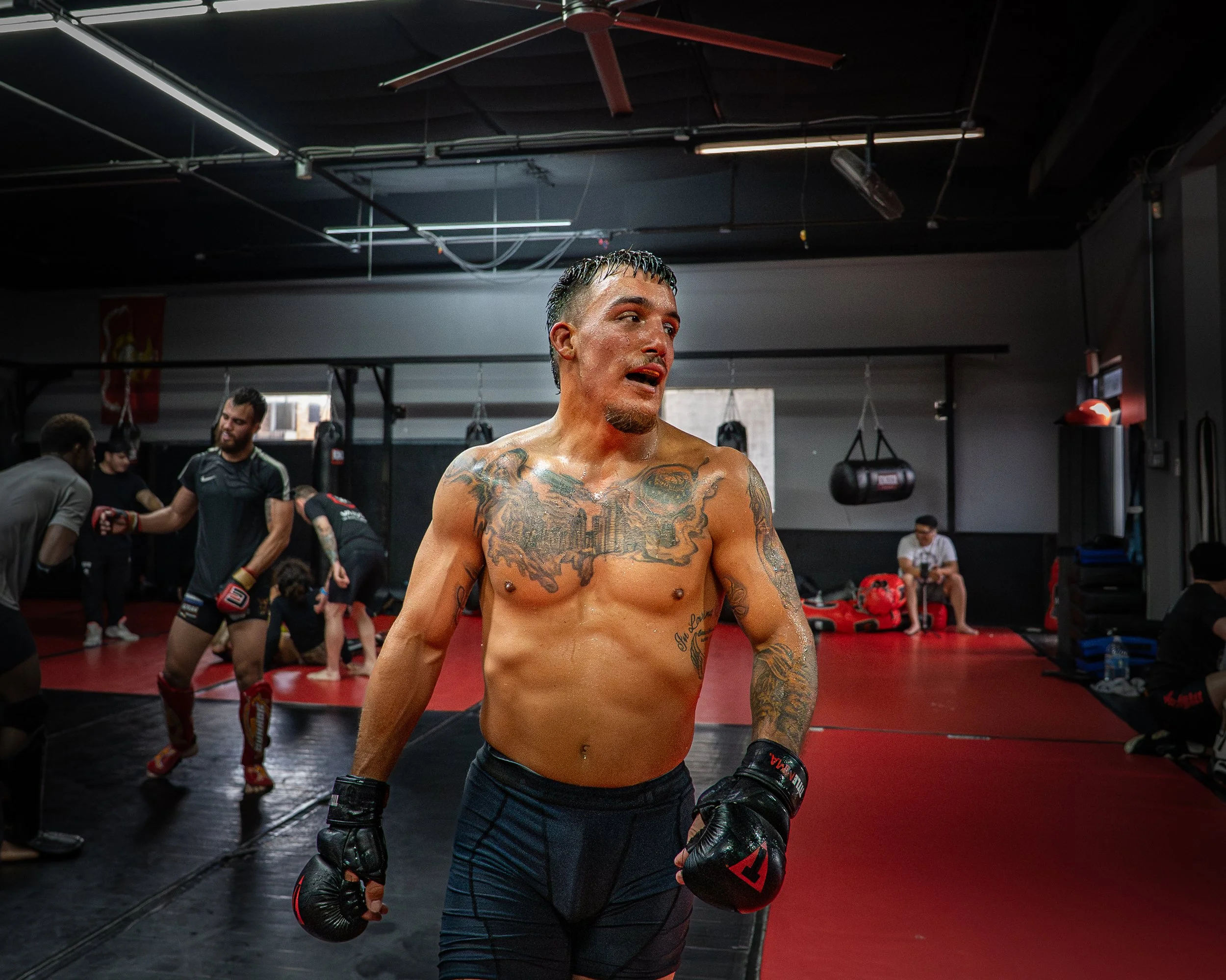 A shirtless male boxer with tattoos standing in a boxing gym, wearing boxing gloves, surrounded by other fighters and trainers, with boxing equipment and punching bags in the background.