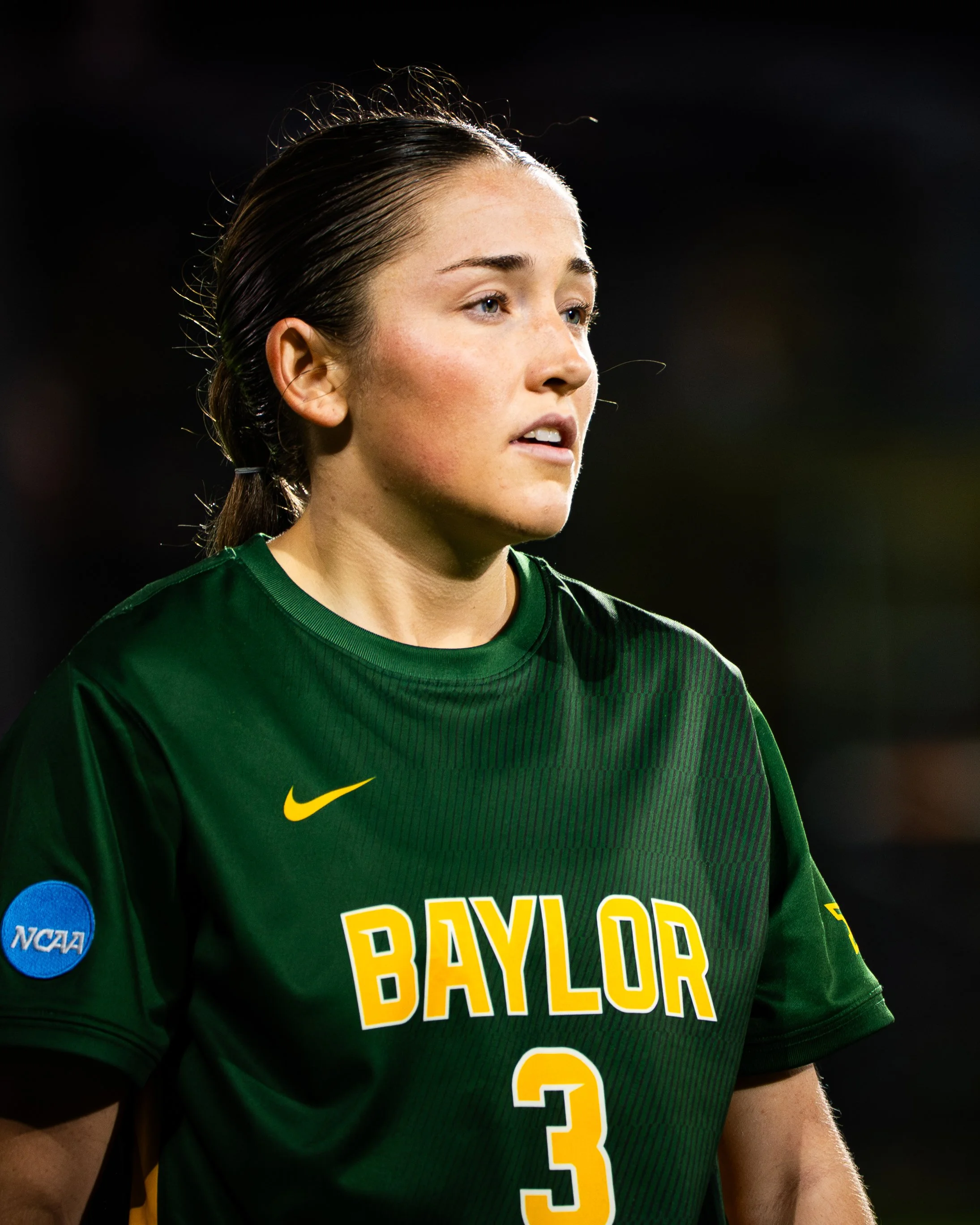 Close-up of a female college basketball player with wet hair, wearing a green Baylor jersey with the number 3, and an NCAA patch on her sleeve, on a dark background.
