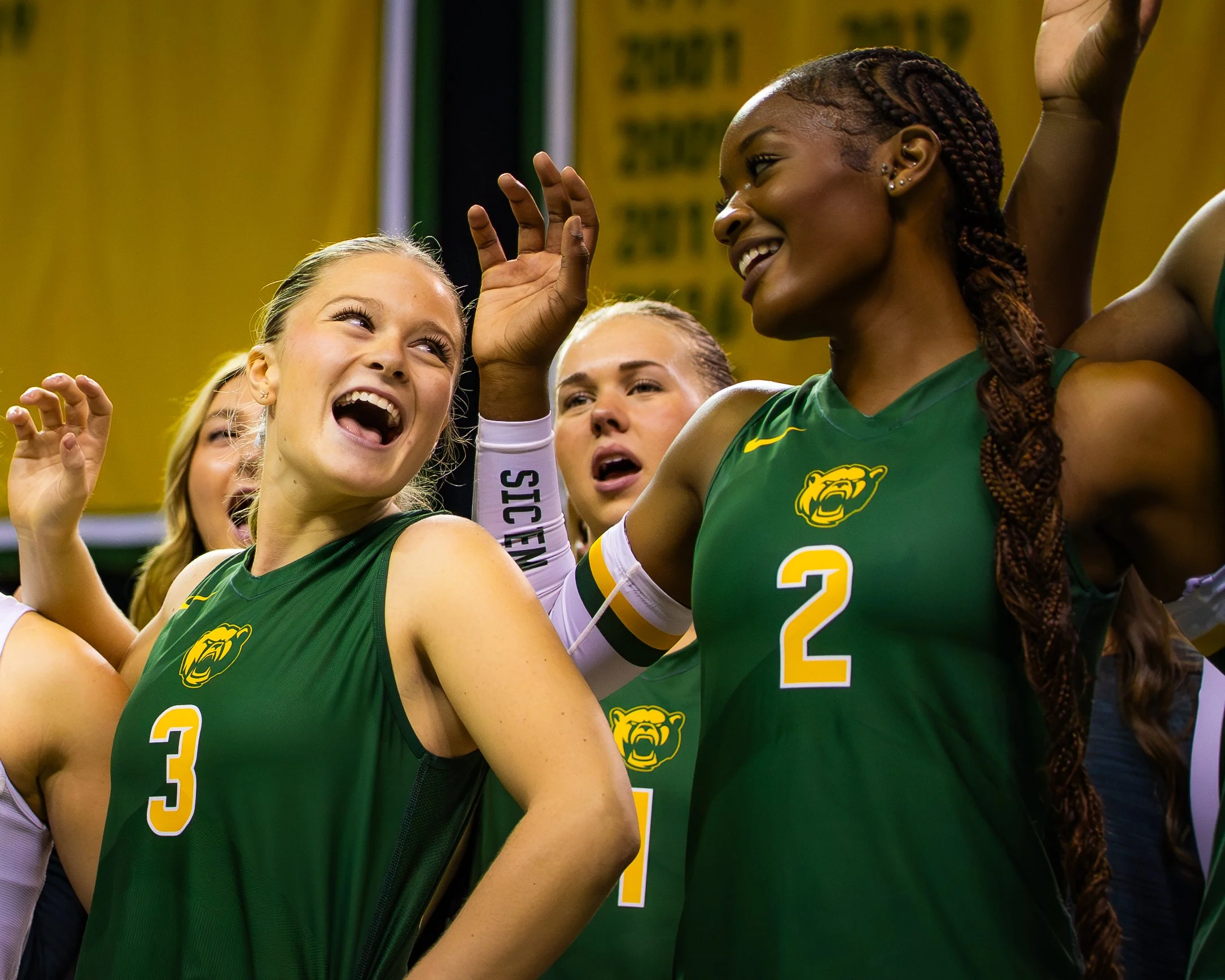 Group of female volleyball players celebrating, wearing green uniforms with a lion logo, in an indoor sports arena.