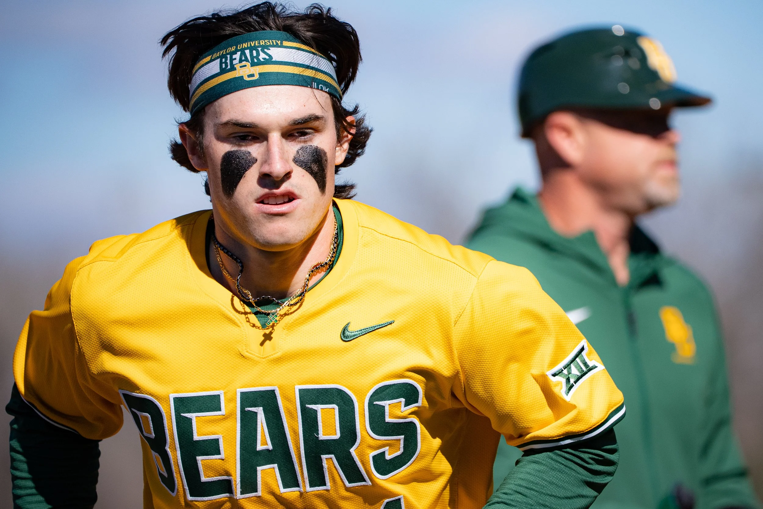 Young man with dark hair wearing a yellow sports jersey and a green and yellow headband with Baylor University Bears logo standing outdoors next to another person in a green and yellow sports uniform.