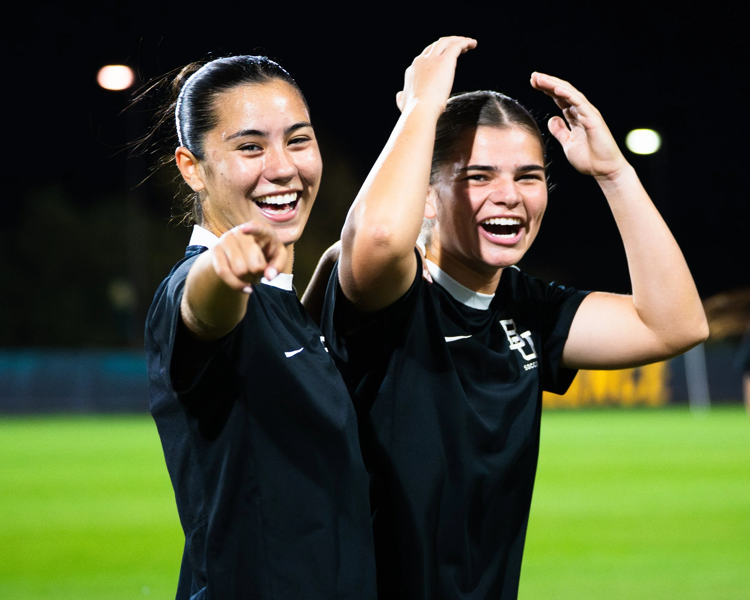 Two young women on a sports field celebrating and smiling, wearing sports uniforms, with a dark background and bright lighting.