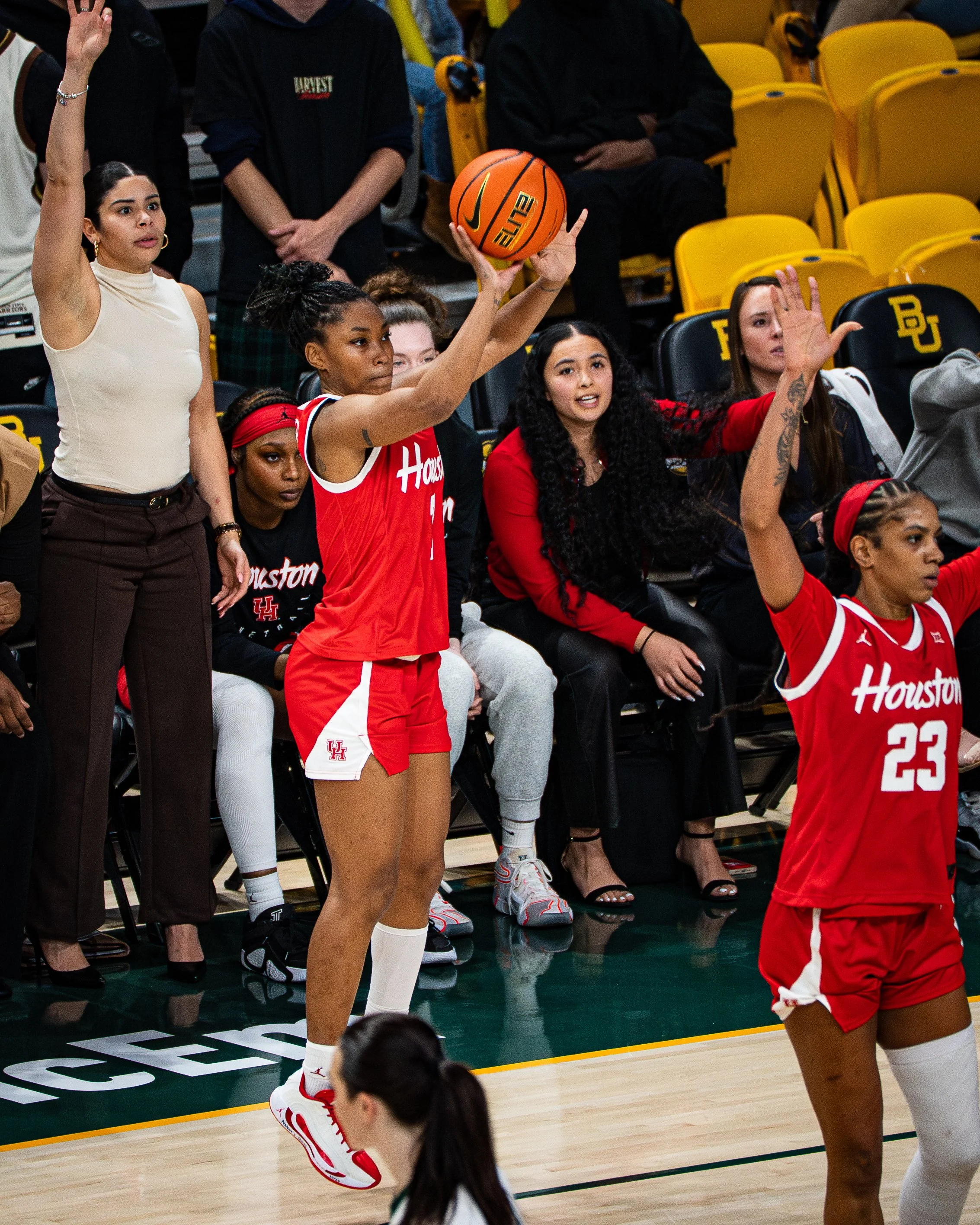 Women on a basketball court, some wearing Houston jerseys, preparing for a game or practice. One woman in a red Houston jersey is holding a basketball, and others are raising their hands or sitting on the sidelines. The background shows empty yellow 