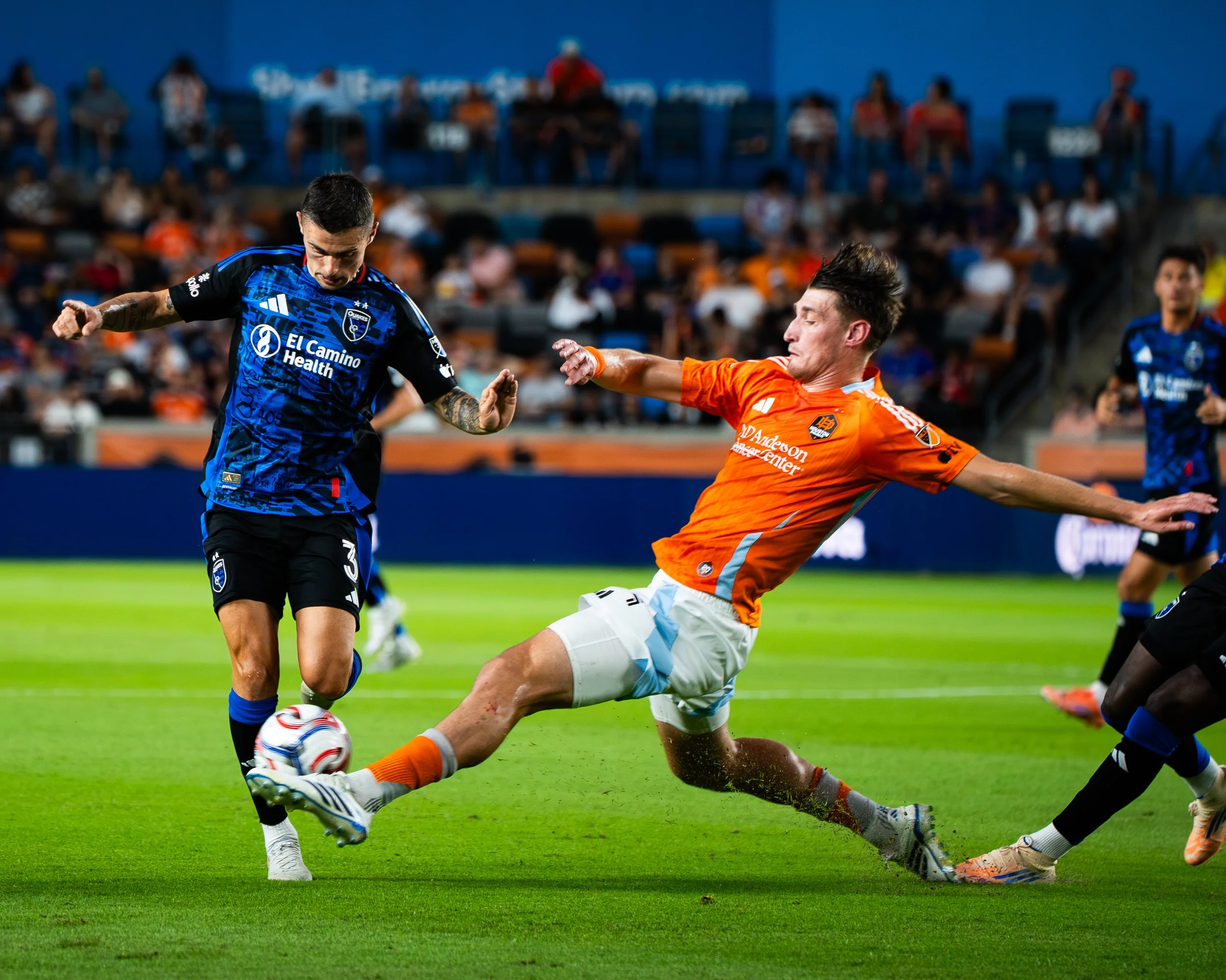 Two soccer players in action on the field, one in a blue uniform and the other in an orange uniform, competing for the ball. The blue team's jersey features the logo of El Camino Healthcare and a team crest, while the orange team's jersey shows the l