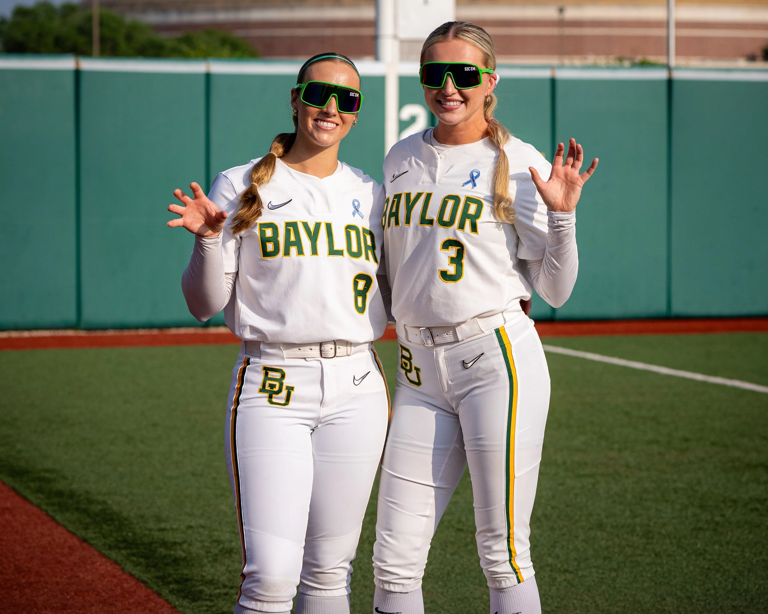 Two female soccer players in white jerseys with 'BAYLOR' written in green and yellow, wearing black and green goggles, smiling and waving at the camera on a soccer field.