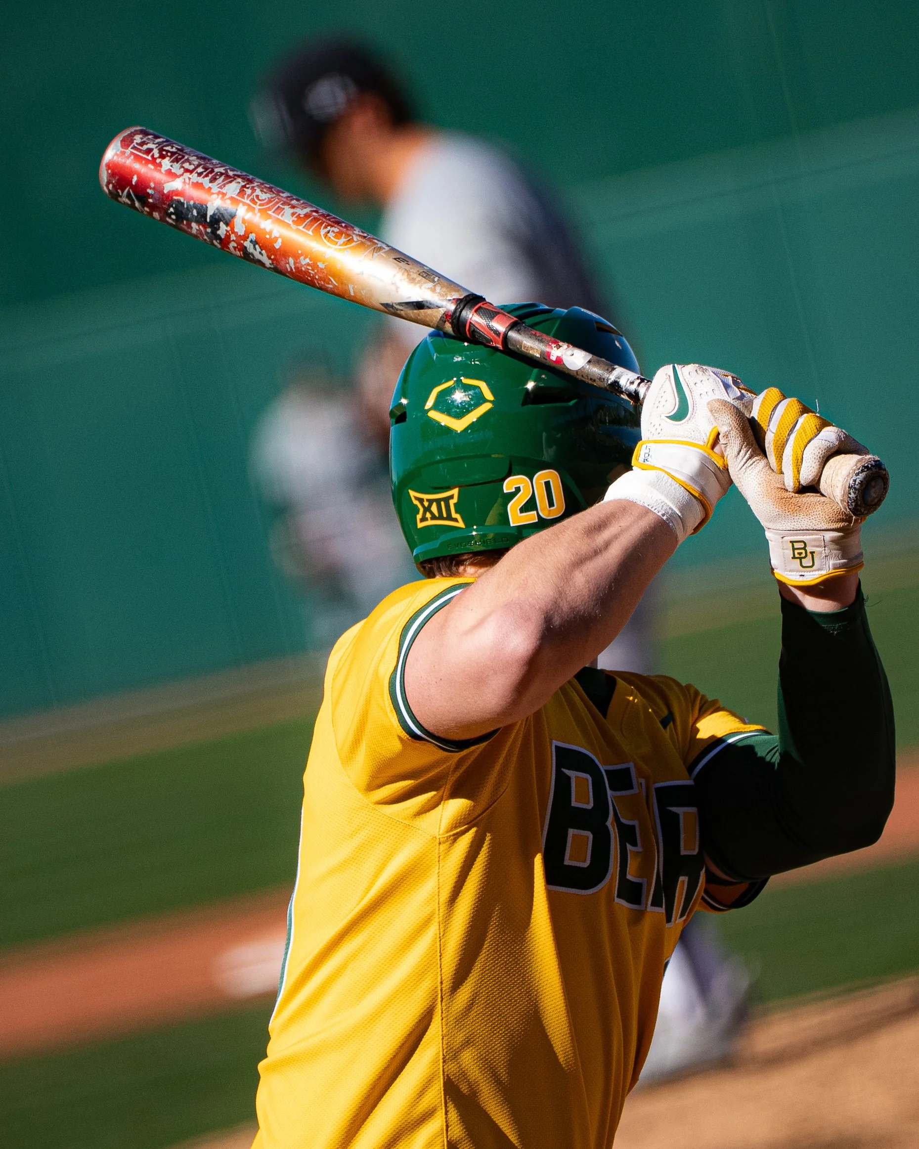 A baseball player in a yellow uniform holding a bat above his head, wearing a green helmet with the Baylor University logo, with a blurred baseball field in the background.