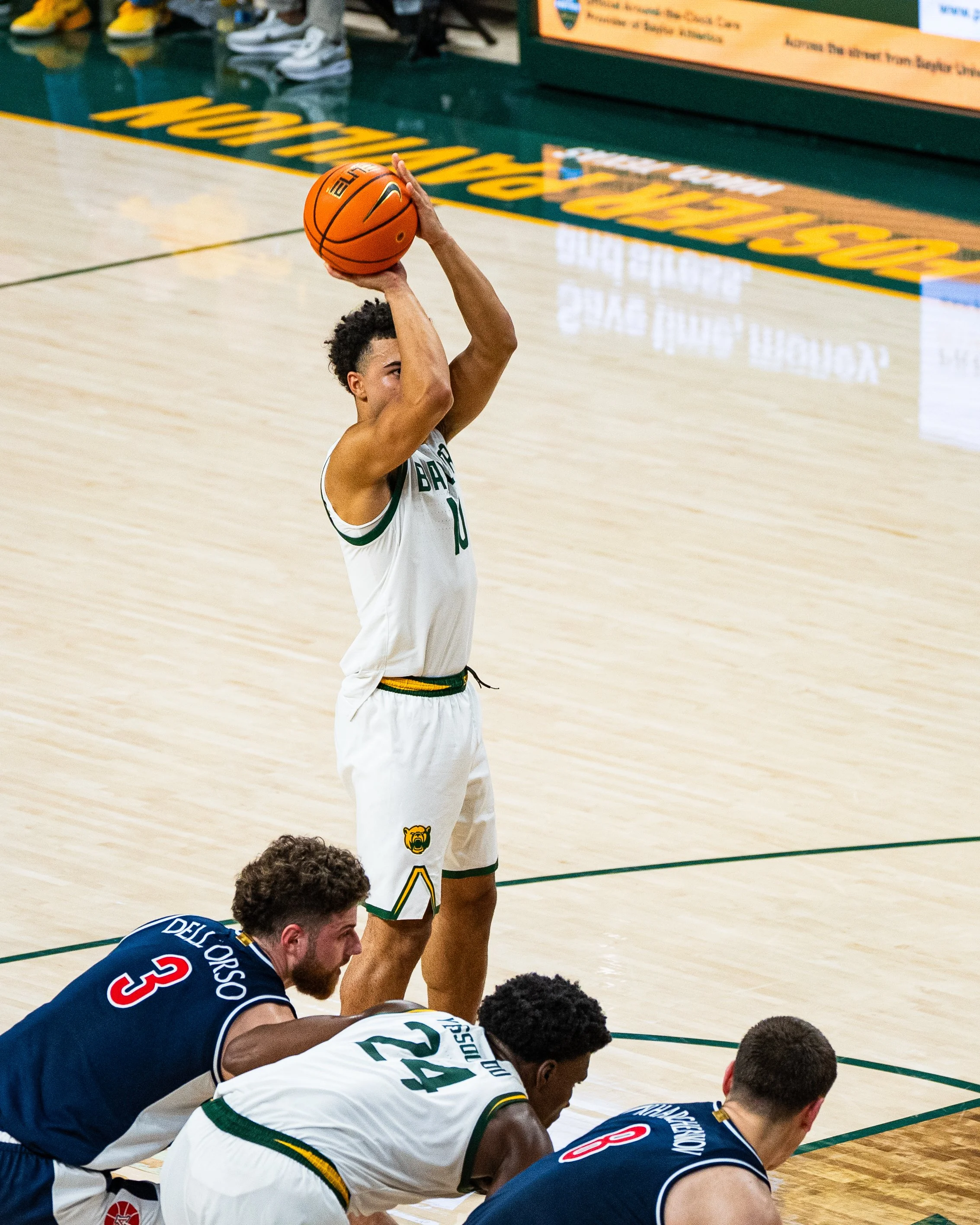 A basketball player prepares to take a free throw shot during a game, wearing a white jersey with green and yellow accents, with teammates and opponents in positions on the court.