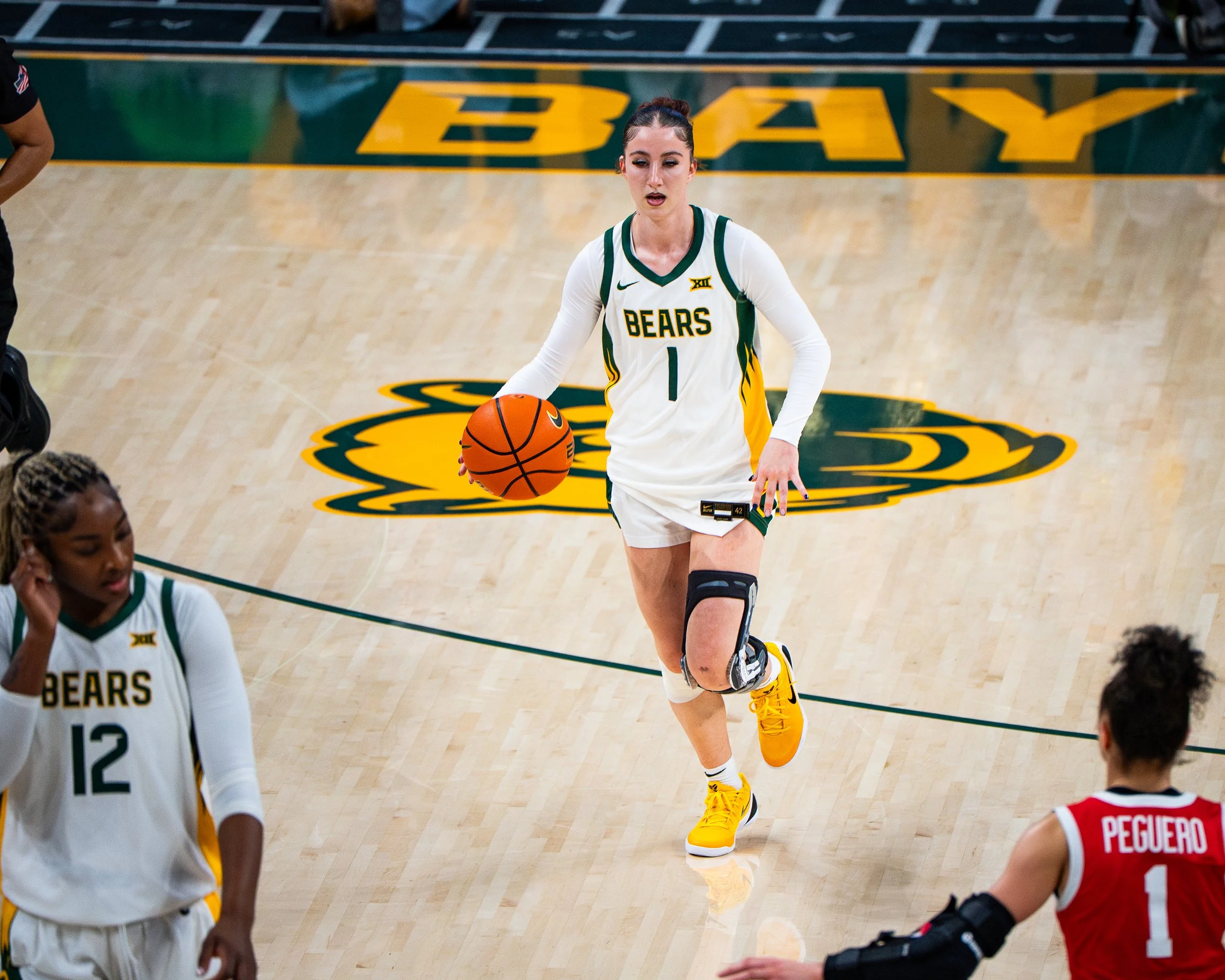 Female basketball player wearing a white and green uniform with yellow shoes dribbling a basketball on a court with a bear logo. Several other players are nearby.