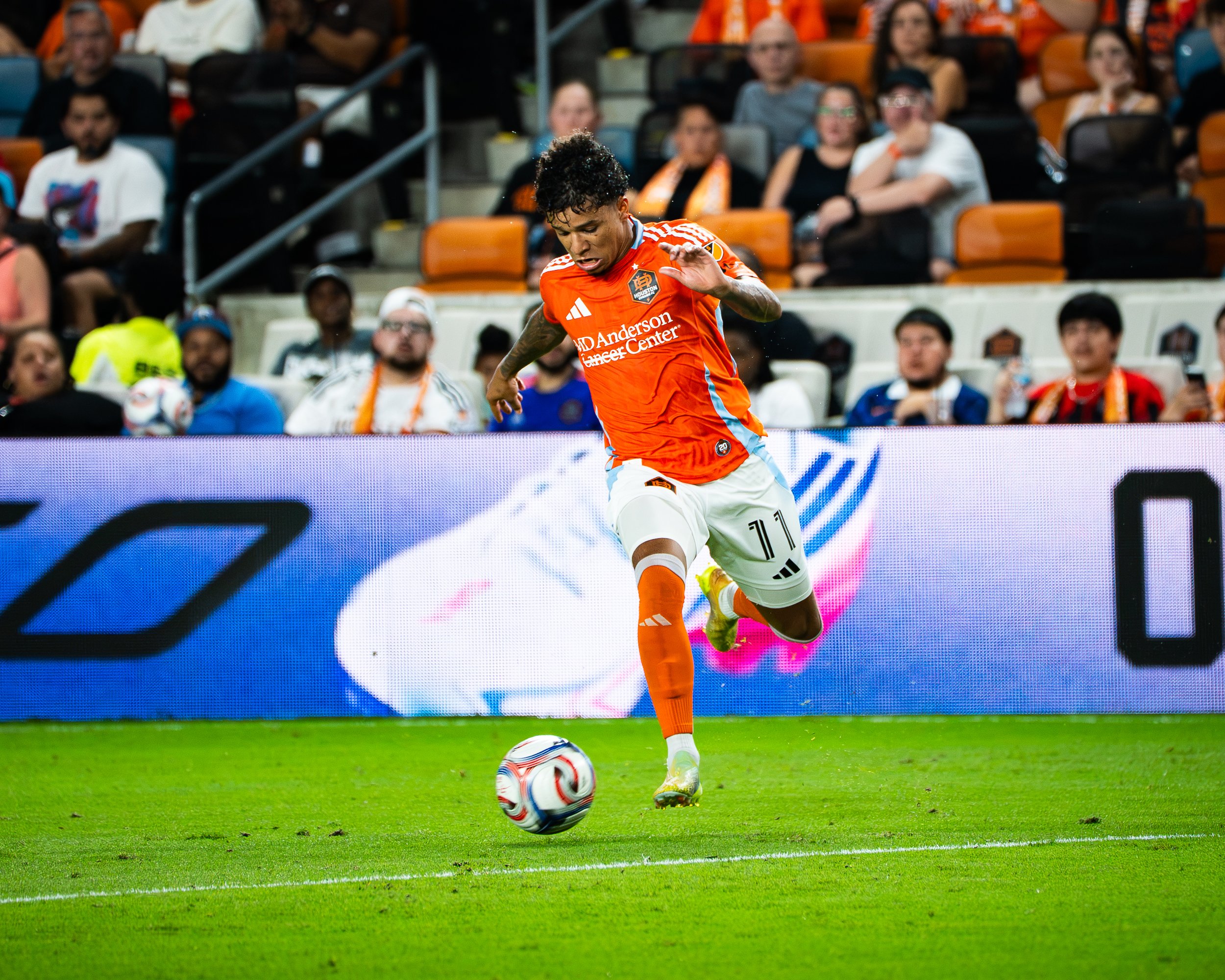 Soccer player in orange uniform kicking a white and red soccer ball during a game, with spectators watching in the background.