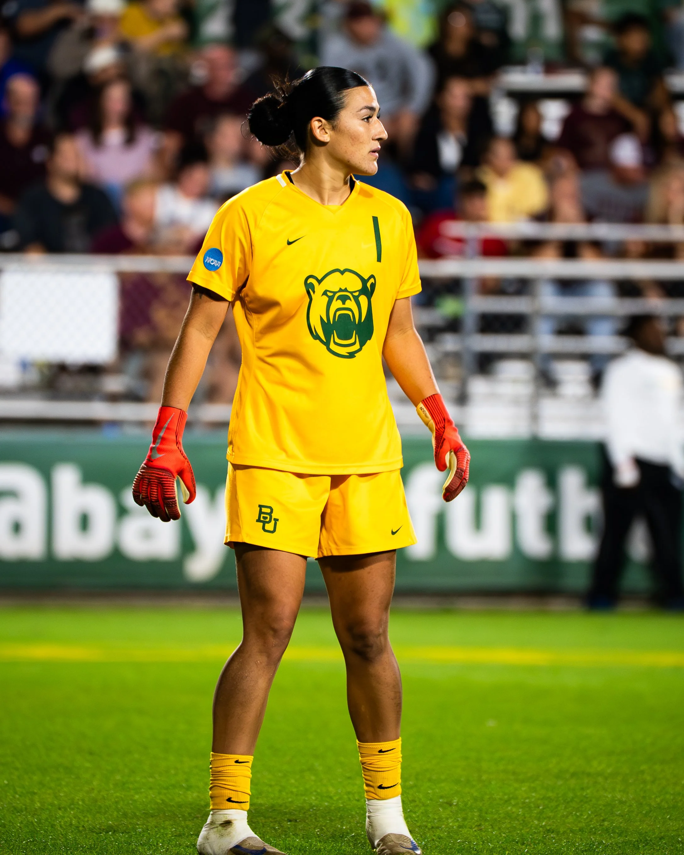 A female soccer player wearing a yellow uniform with a bulldog emblem and a green number 1, standing on a green field during a game, with a blurred crowd in the background.