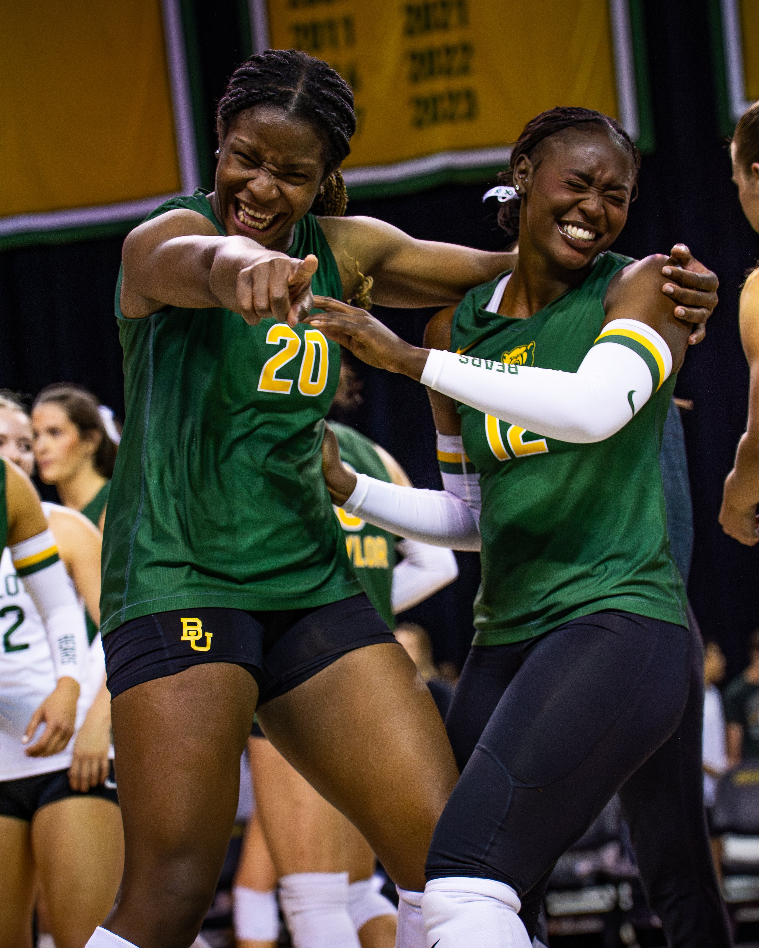 Two female volleyball players from Baylor University celebrating during a game. They are wearing green jerseys with yellow numbers, and one is pointing while the other is smiling with eyes closed.