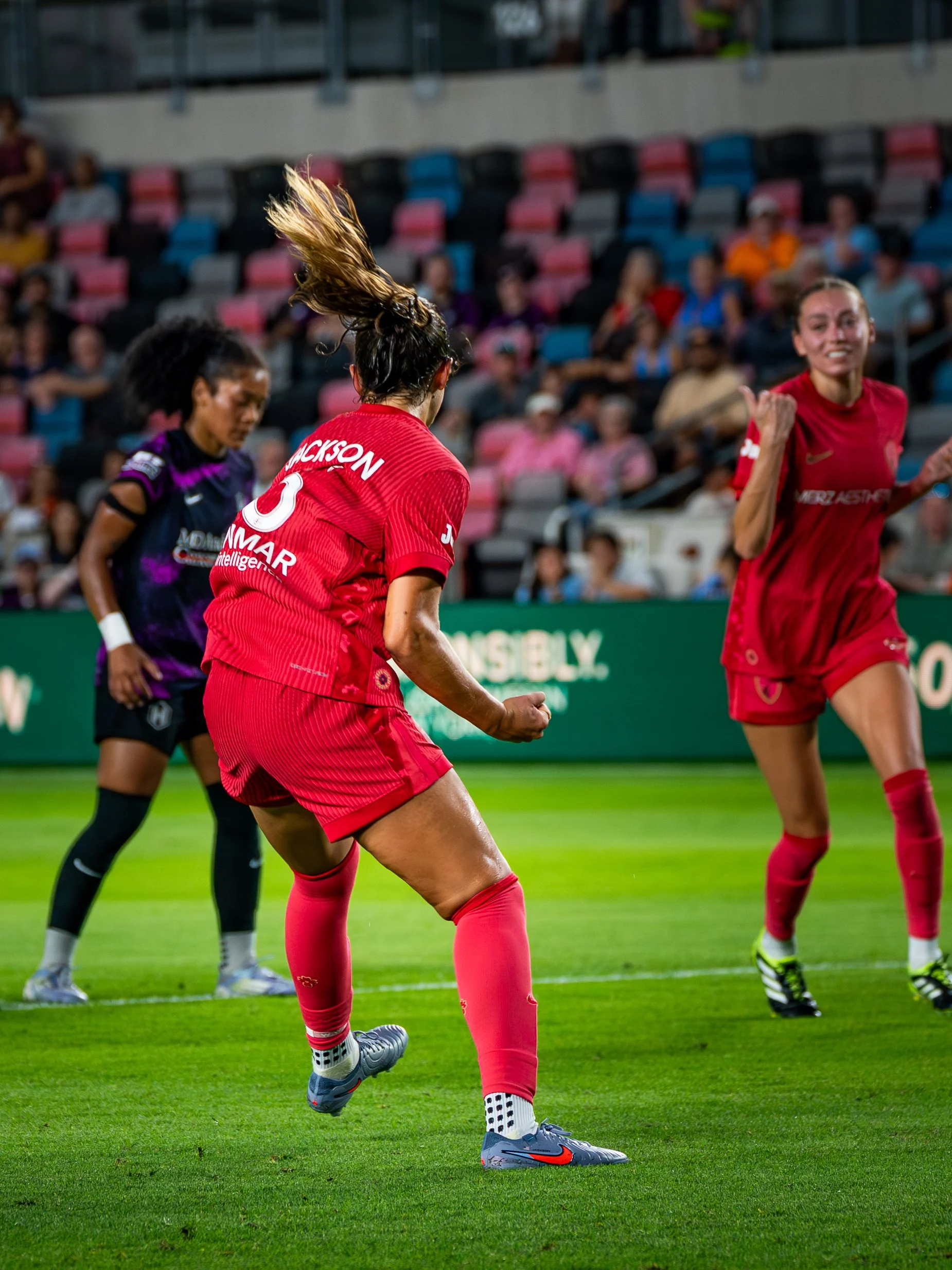 Women playing soccer on a field, two wearing red jerseys and one in a purple and black jersey, with spectators in the stands.