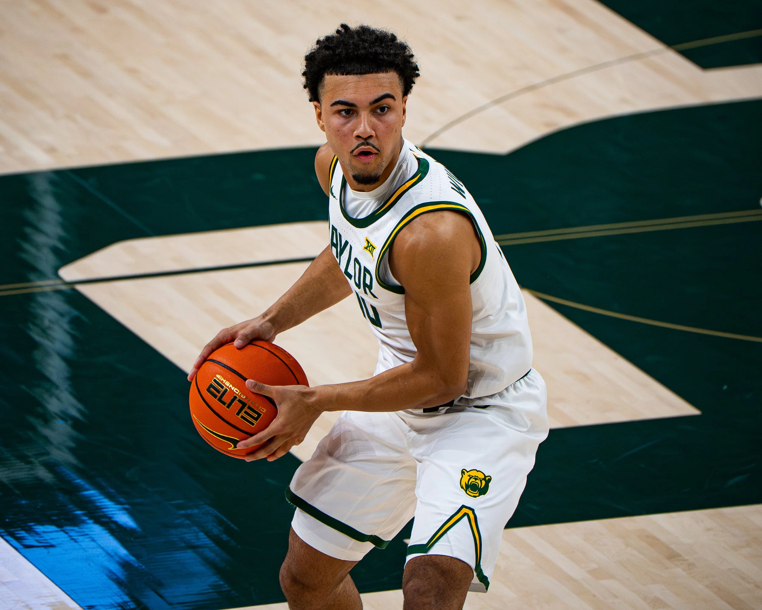 A basketball player in a white jersey with yellow and green accents dribbling the ball on a basketball court. The player's jersey has a bear paw logo and the words "WILDCATS".