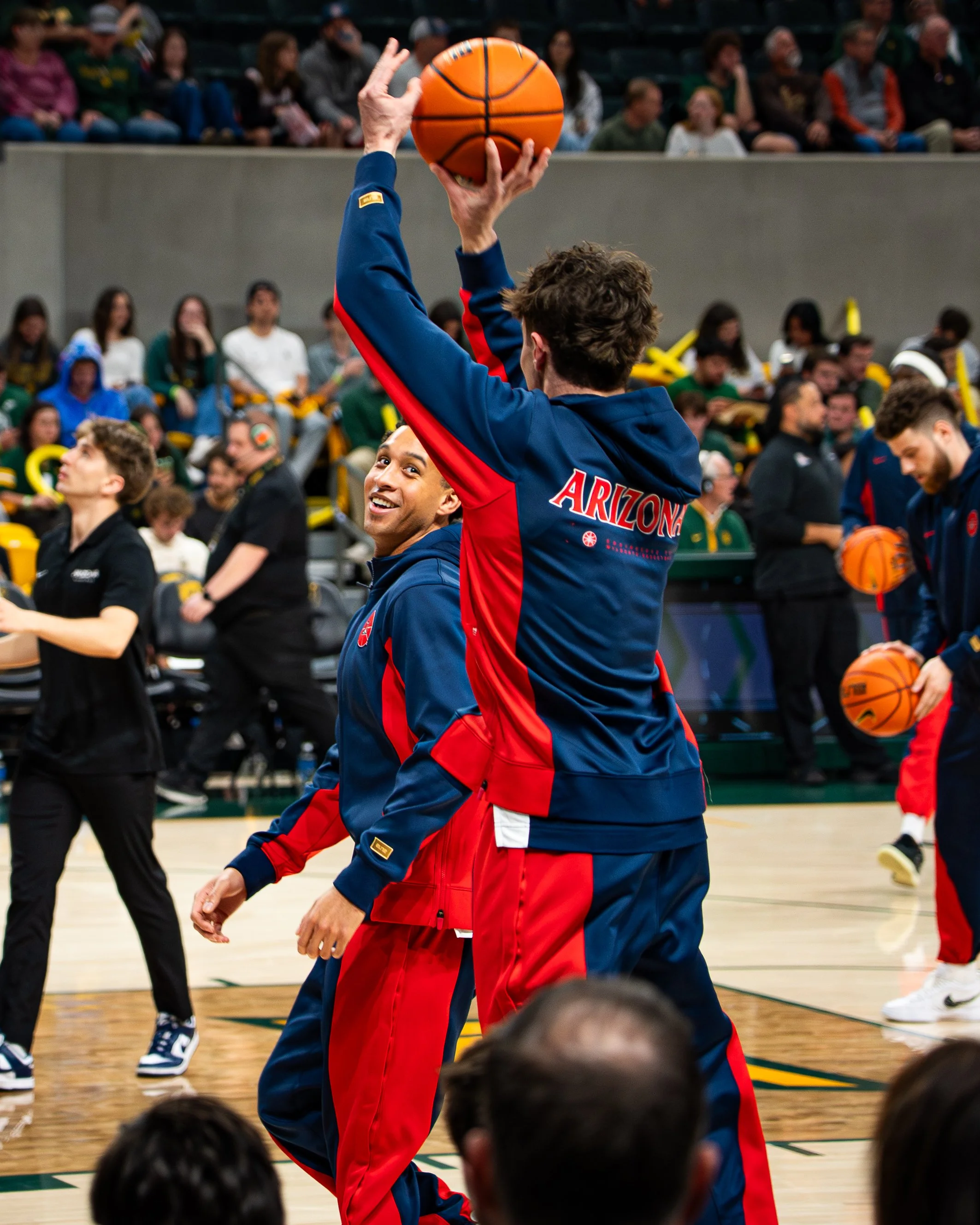 A basketball player wearing a jacket with 'ARIZONA' on the back is holding a basketball above his head on an indoor basketball court during a game or practice, with spectators seated in the background.