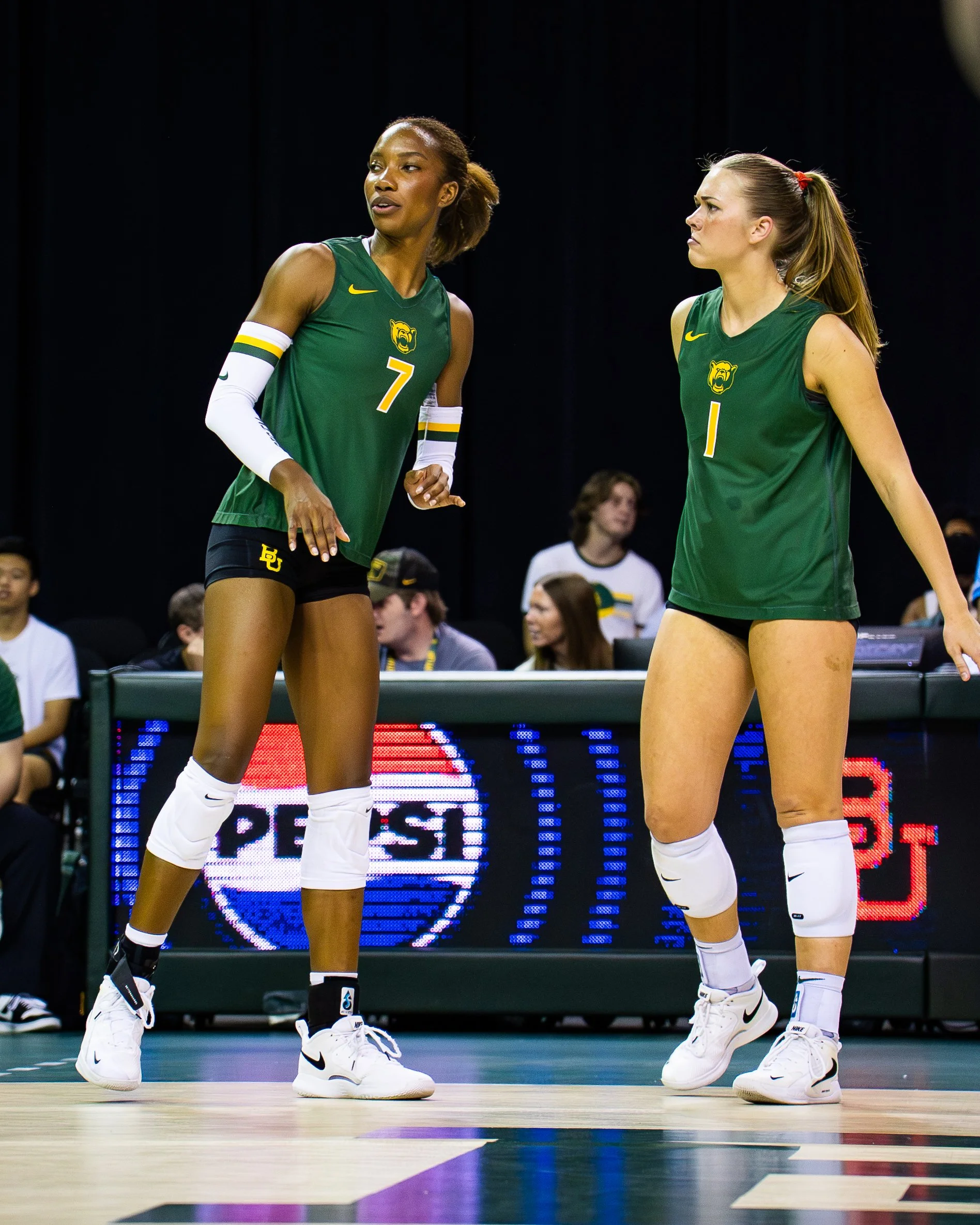 Two female basketball players in green and yellow uniforms standing on a basketball court, engaged in a conversation during a game or practice.