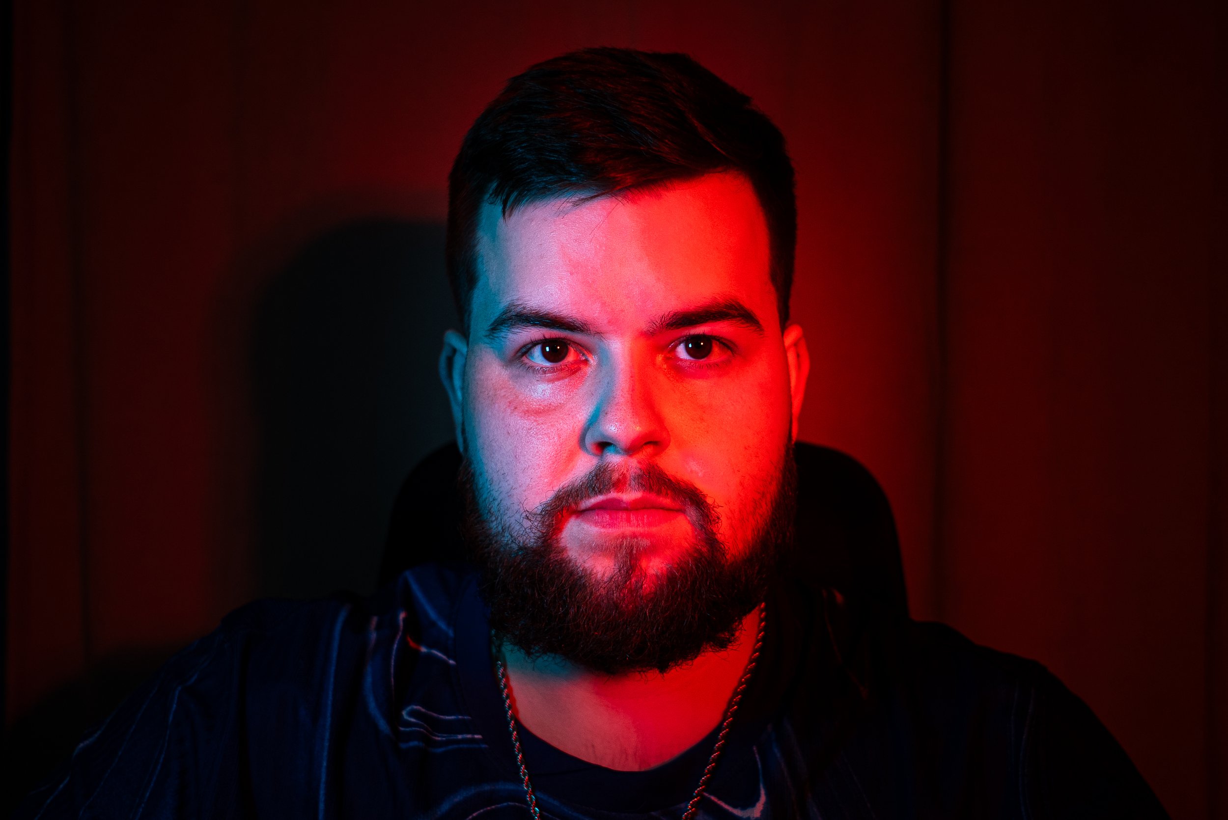Close-up portrait of a man with dark hair and beard, illuminated with red and blue lighting, wearing a dark jacket with pinstripes and a chain necklace.