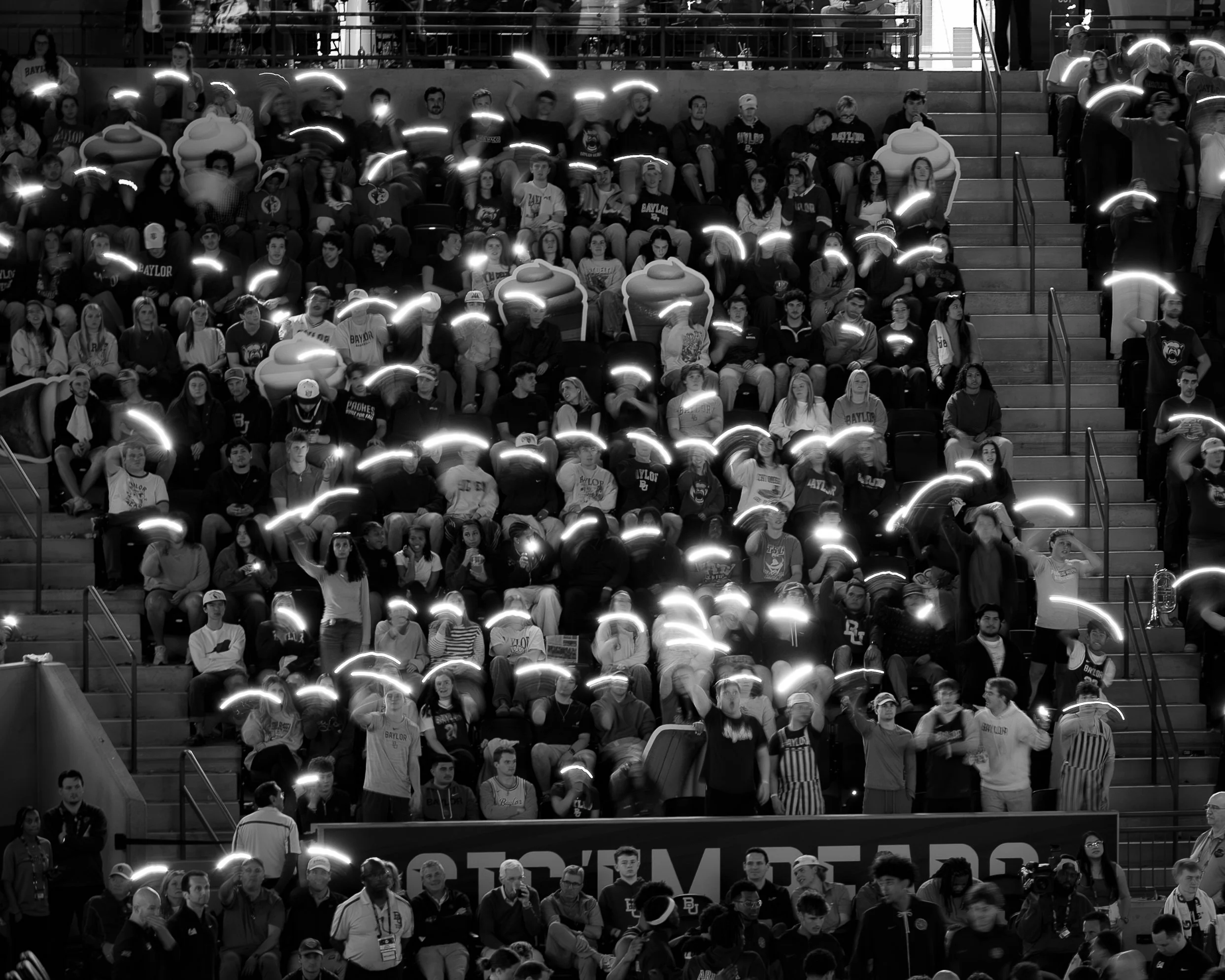 A black and white photo of a large group of people seated in stadium bleachers. Some are holding illuminated objects, creating light trails above their heads. The crowd appears to be attending a sports event or concert.