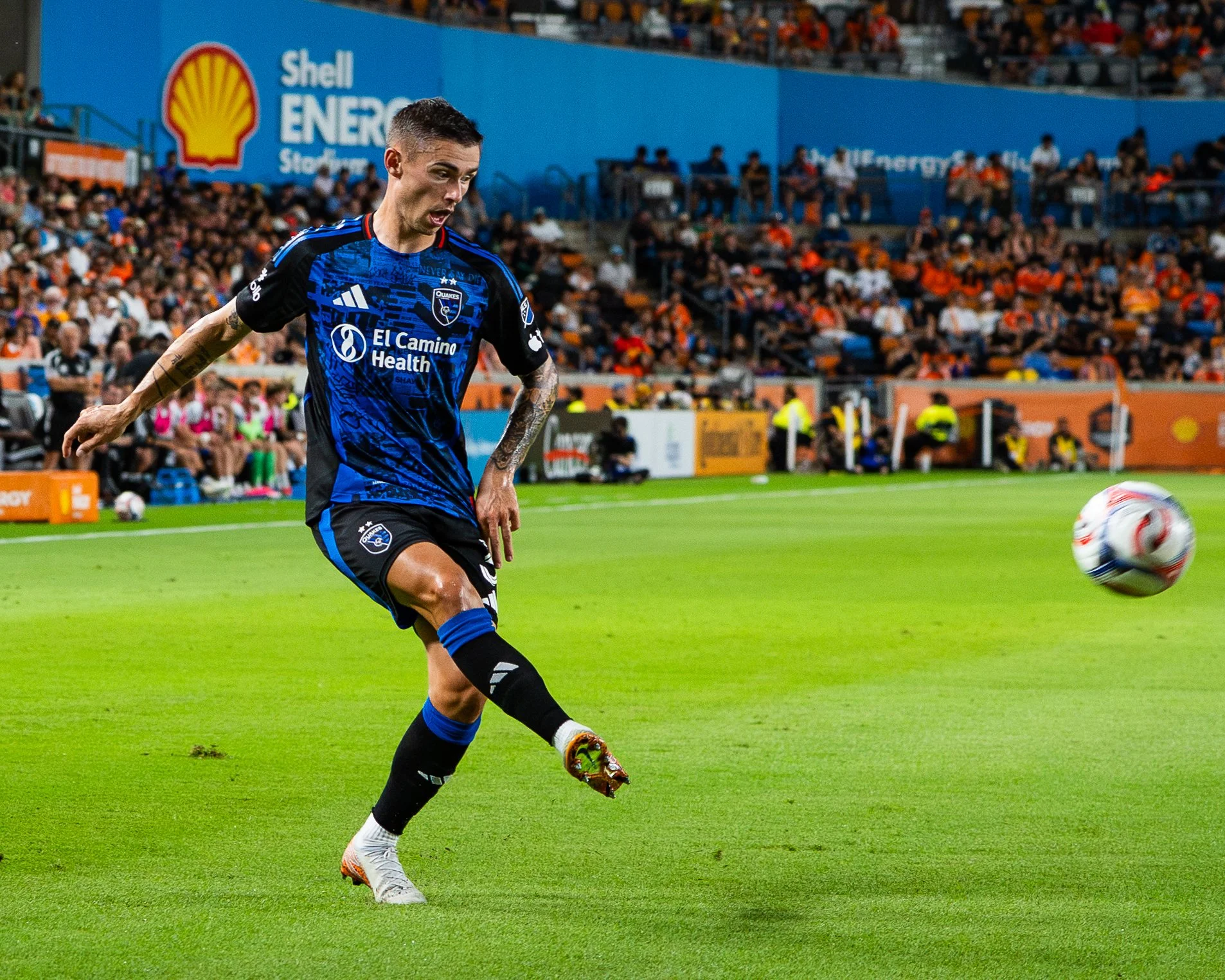 A soccer player in a blue and black uniform with tattoos on his arms, is kicking a soccer ball on a green field during a game. In the background, there are spectators in the stands and security personnel near the sidelines.