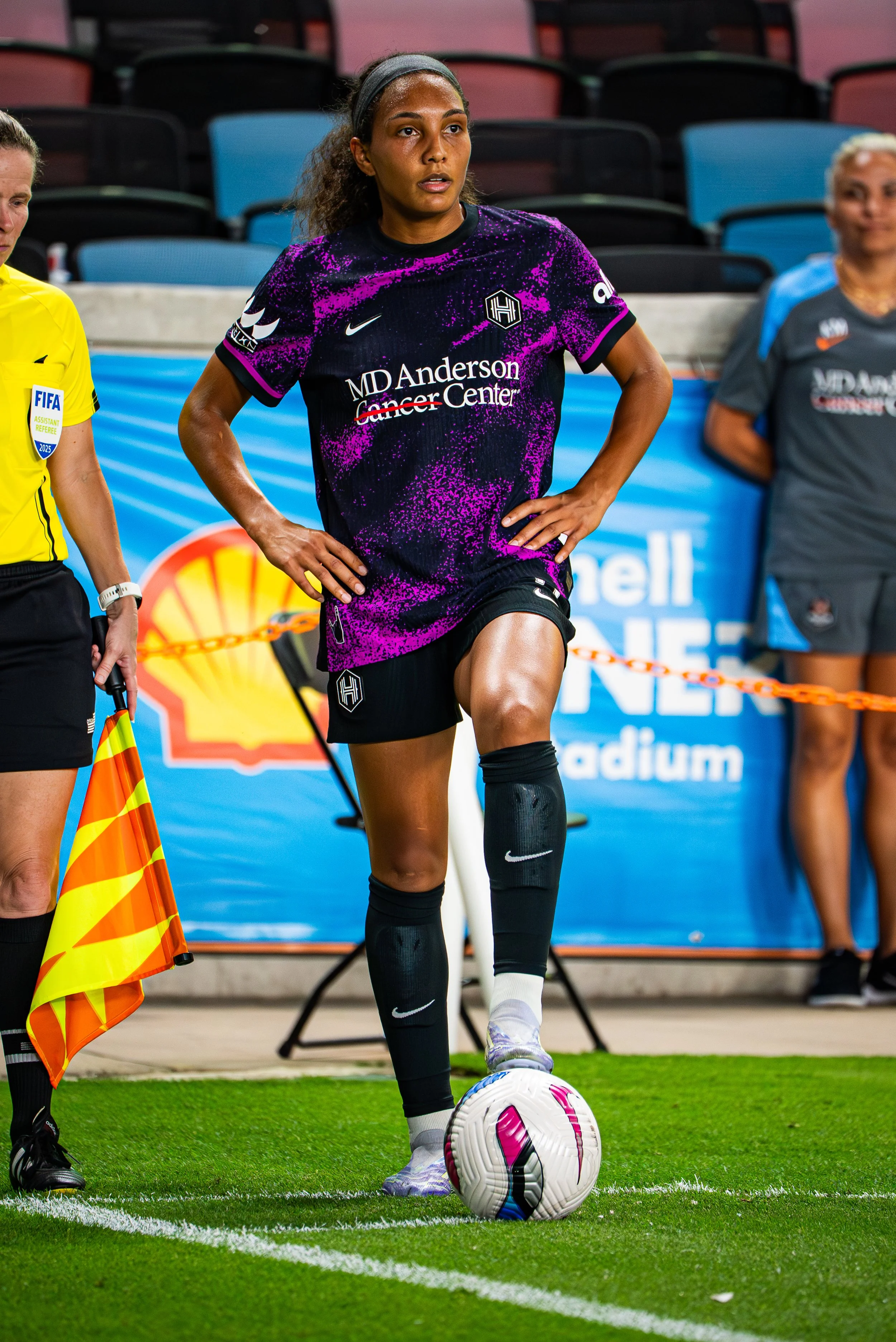 A female soccer player in a purple and black uniform preparing to kick a soccer ball during a game, with a referee and another person in the background.
