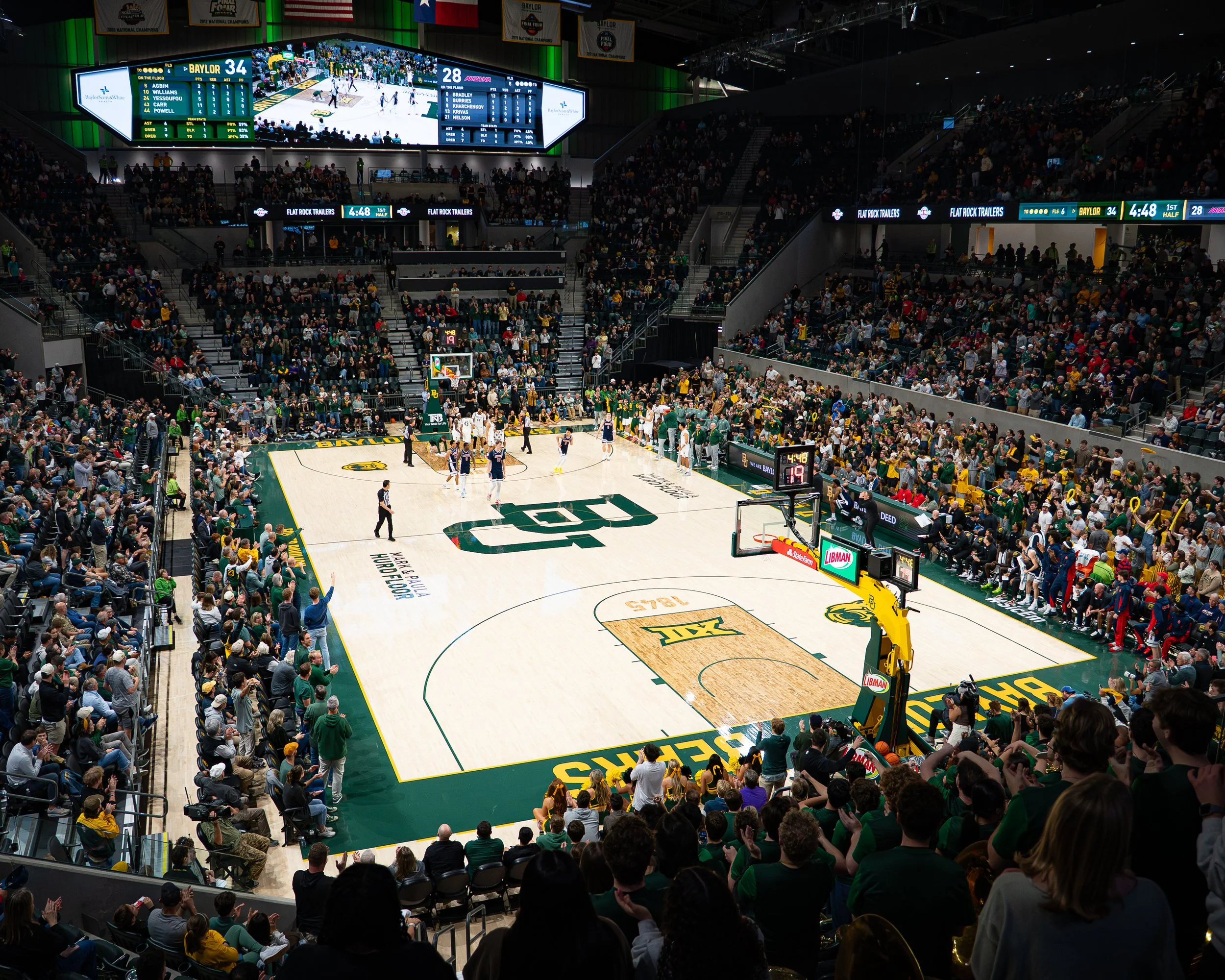 Interior of a basketball arena filled with spectators watching a game. The court is labeled with the University of Texas at Austin logo, and players are on the court. Digital scoreboards and advertisements are visible above the court, displaying the 