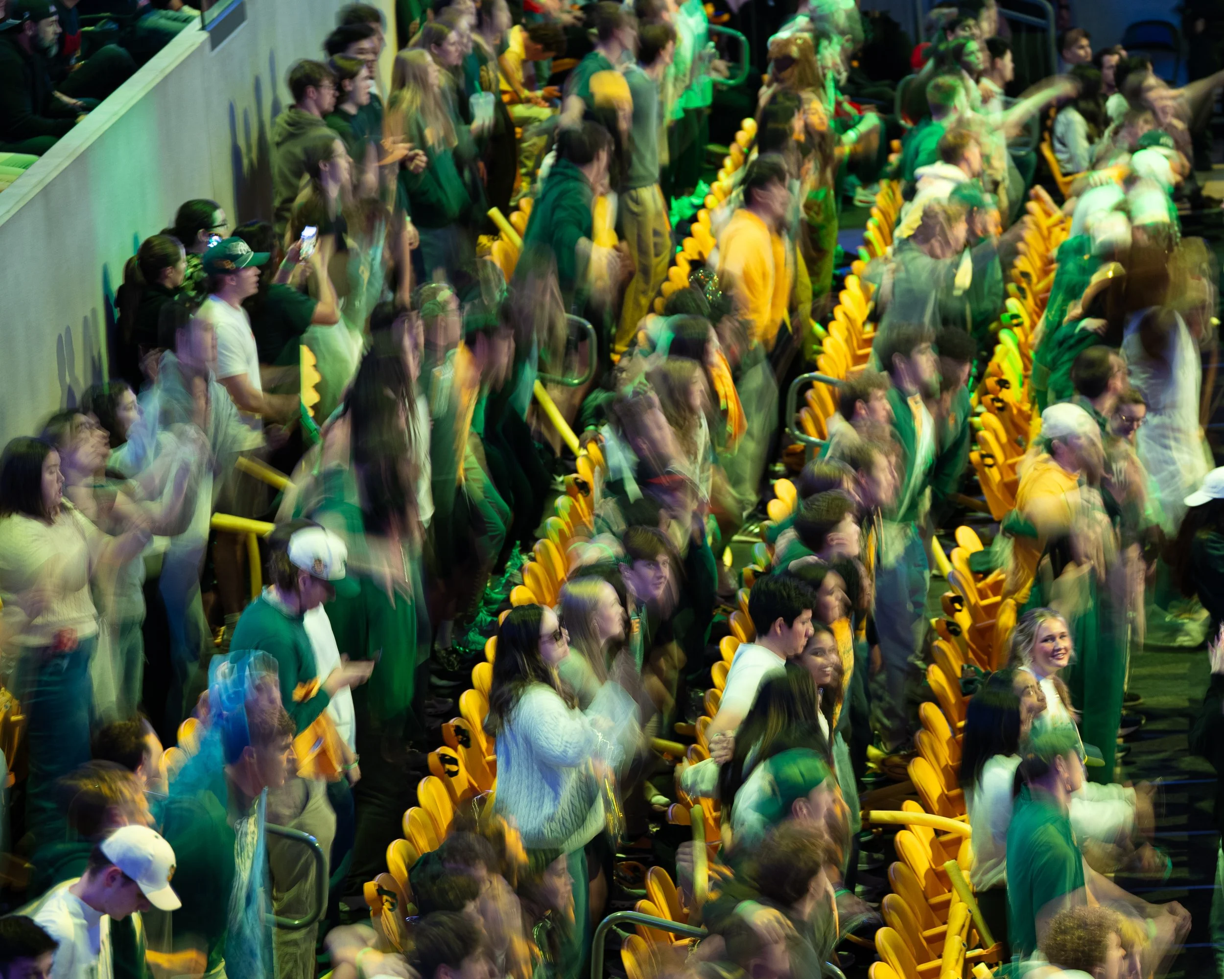 Crowd of people standing and sitting in stadium seats, some taking photos, with yellow seats and green lighting effects.