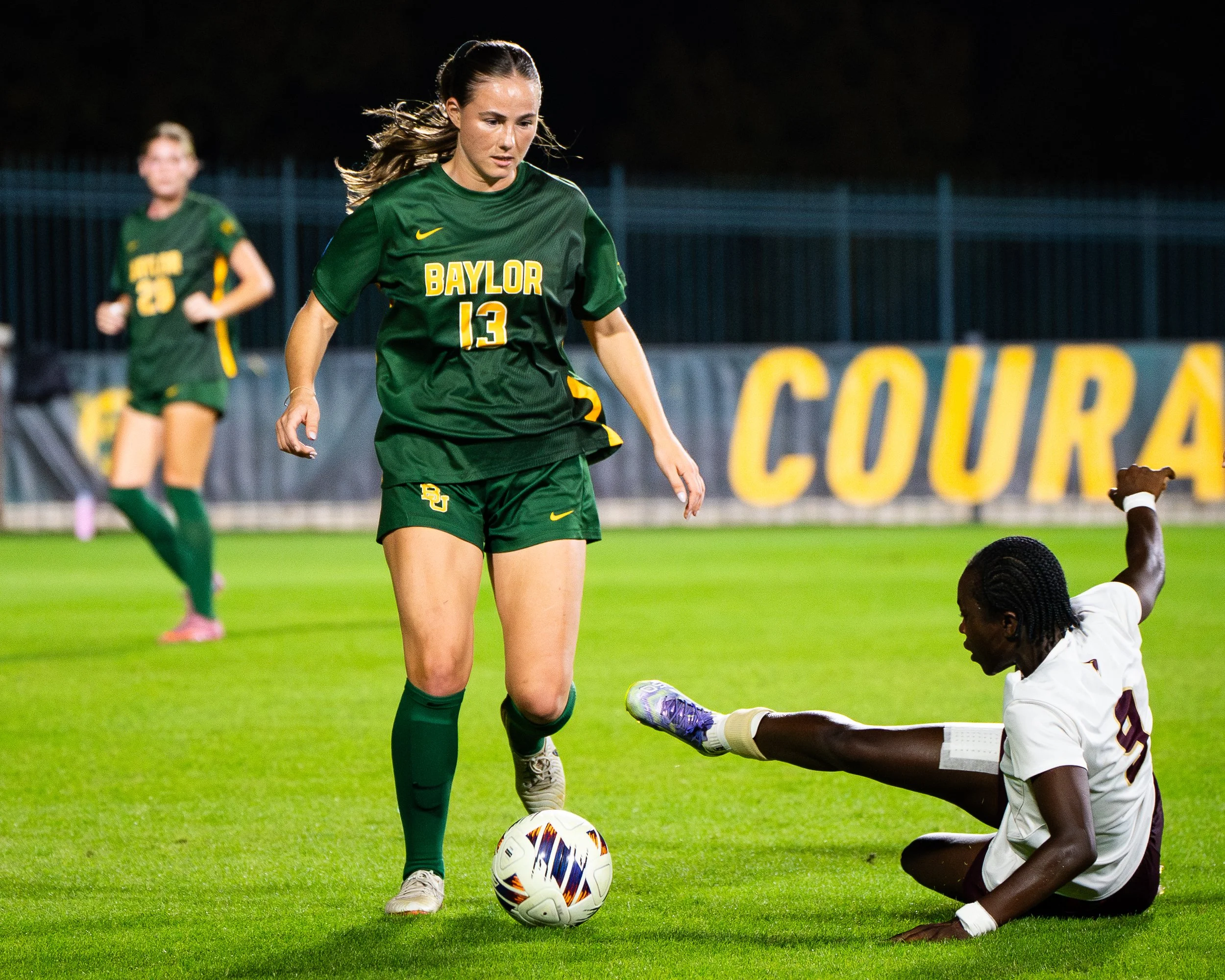 A female soccer player wearing a green Baylor jersey with the number 13, standing on a field next to a fallen opponent. The opponent is on the ground, wearing a white uniform, with one leg extended and an arm raised. Another player in green is blurre