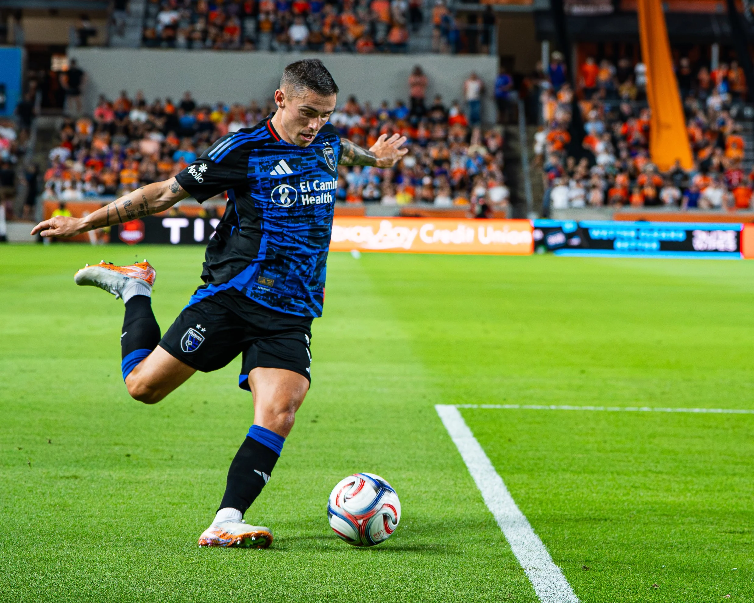 A soccer player in a blue and black uniform kicking a soccer ball on a green field, with fans in the stands in the background.