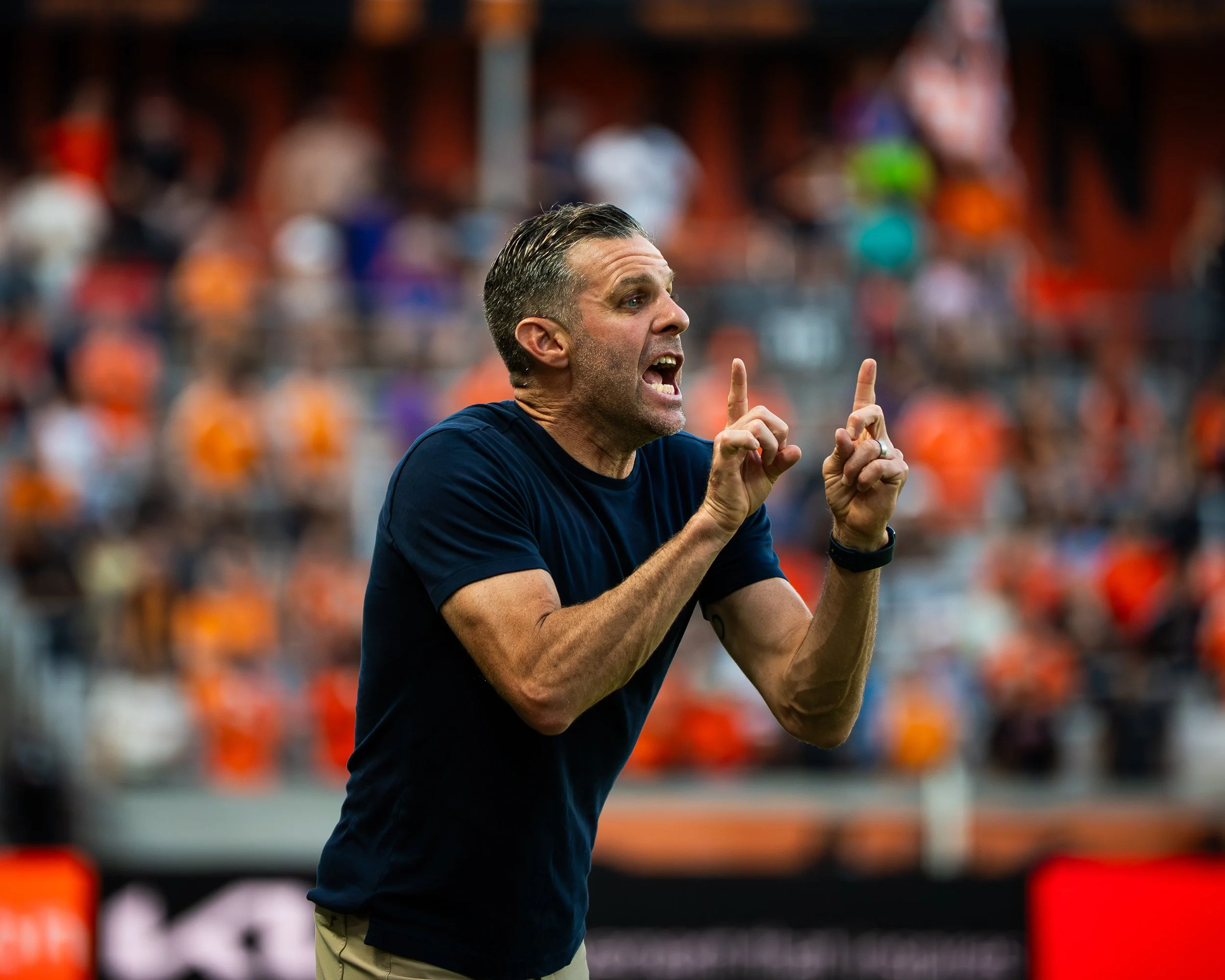 A man with short dark hair, wearing a navy shirt and tan pants, is gesturing with both index fingers raised while talking passionately on a sports field with stadium seats in the background.