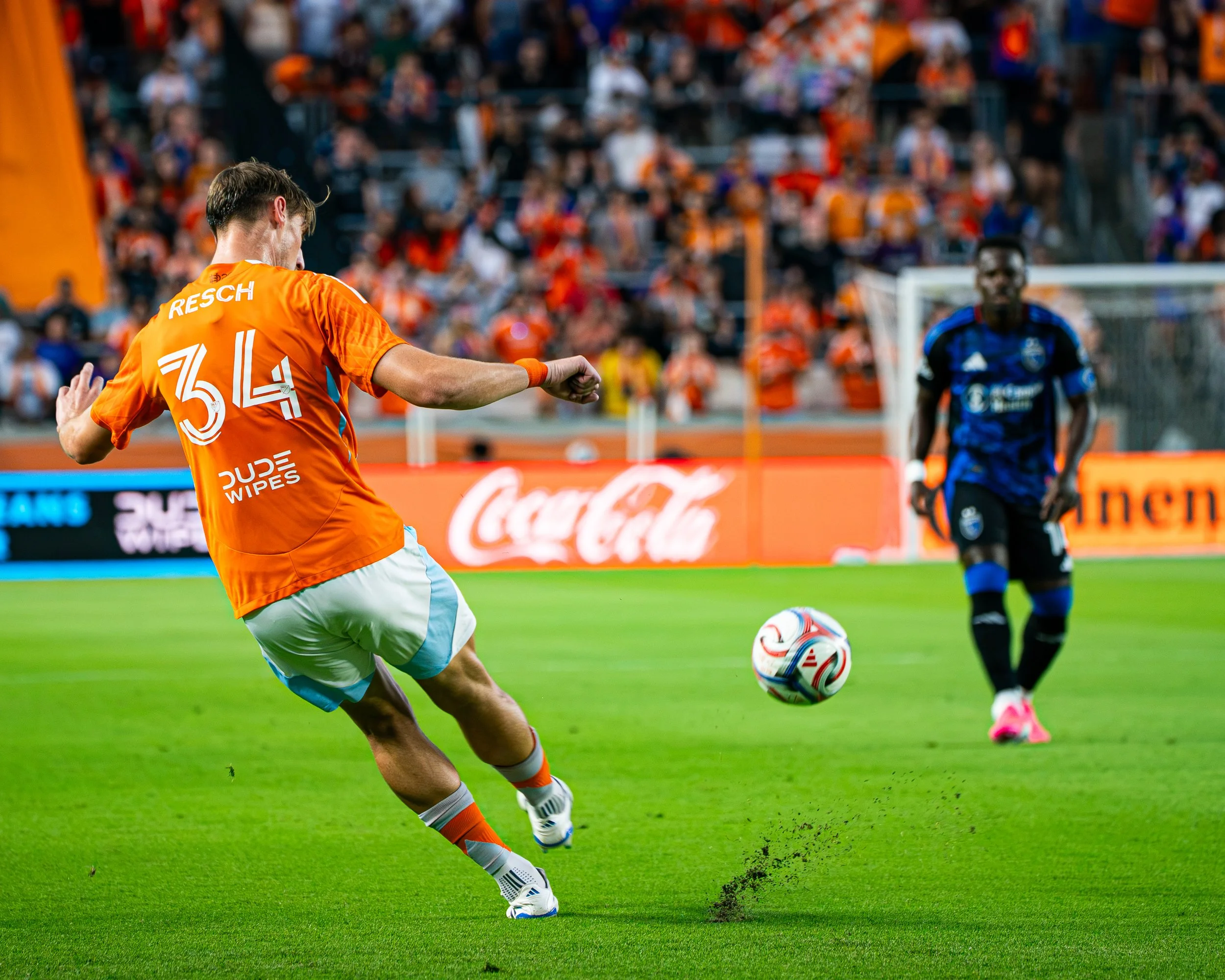 A soccer player in an orange jersey kicking a soccer ball on a green field during a game, with an opponent and spectators in the background.