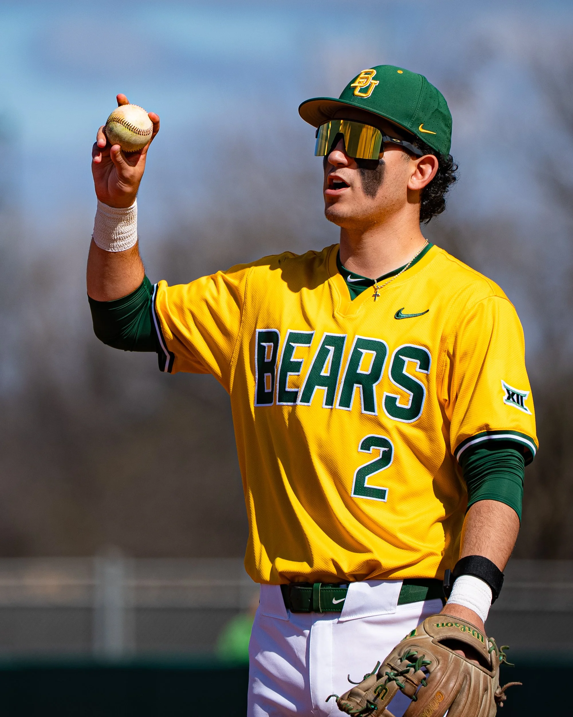 A baseball player in a yellow jersey with 'BEARS' written in green and white letters, holding a baseball in his right hand, wearing a green cap with 'BU' on it, black sunglasses, and a beige glove on his left hand.