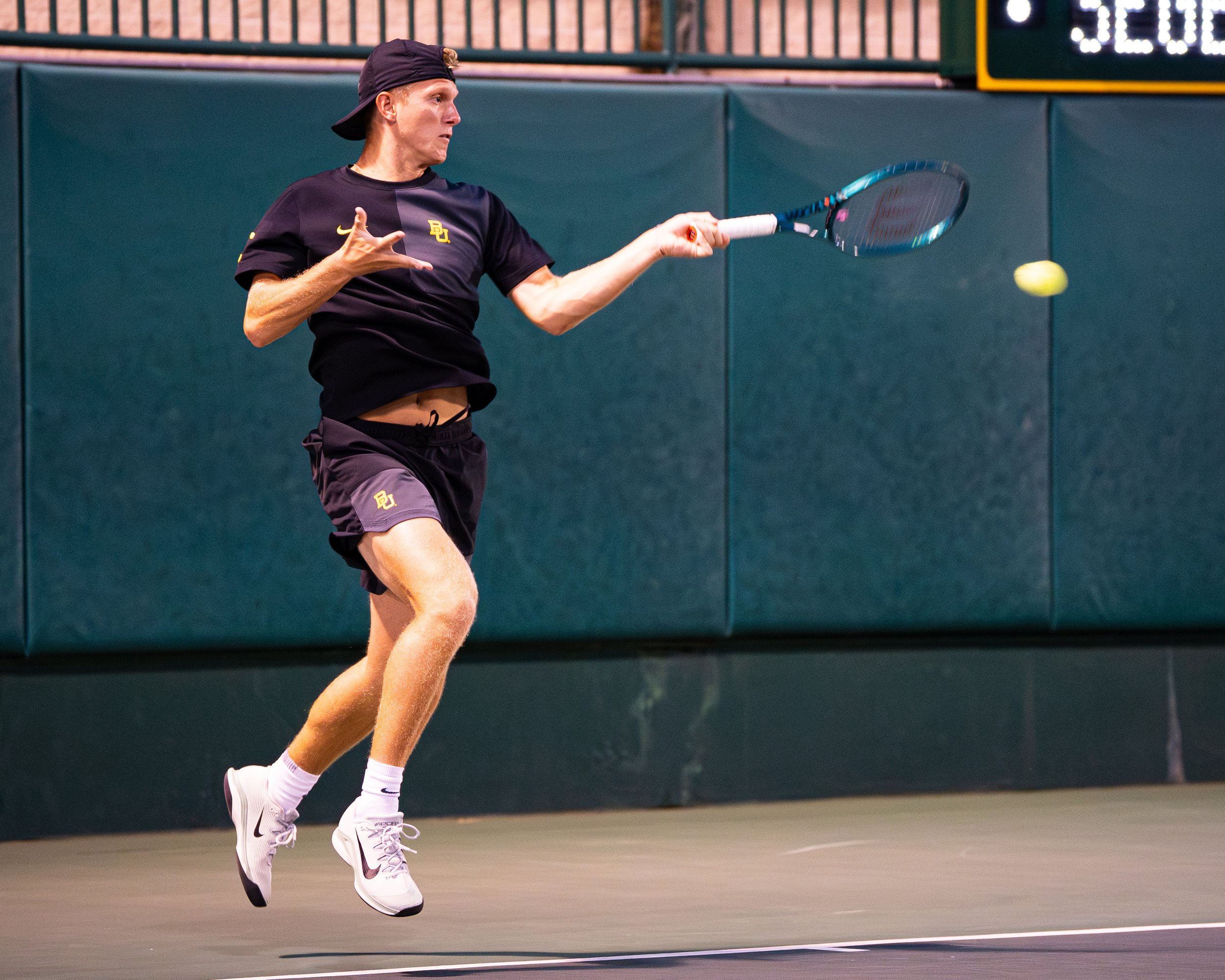 A male tennis player wearing a black t-shirt, black shorts, white Nike tennis shoes, and a black cap, playing tennis on an indoor court.