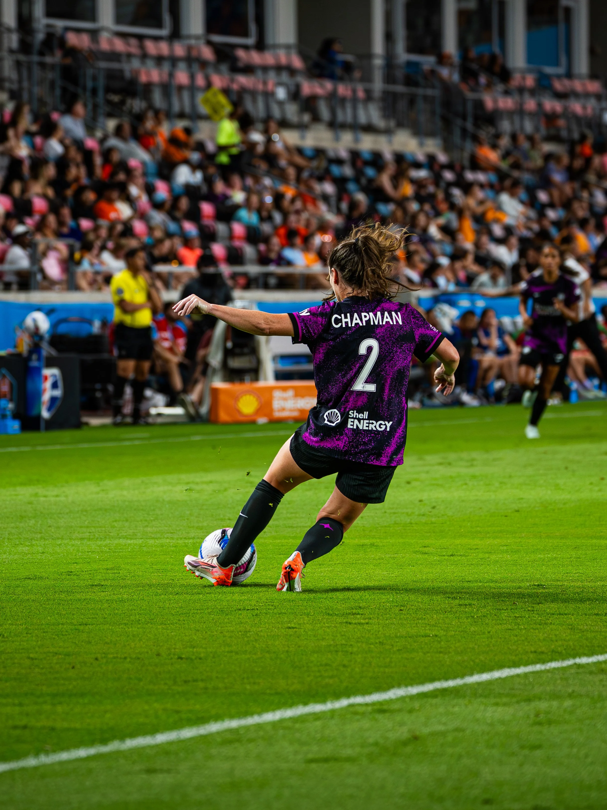 A female soccer player wearing a black and purple jersey with the name 'Chapman' and the number 2 is kicking a soccer ball on a green field. There are spectators in the stands in the background.