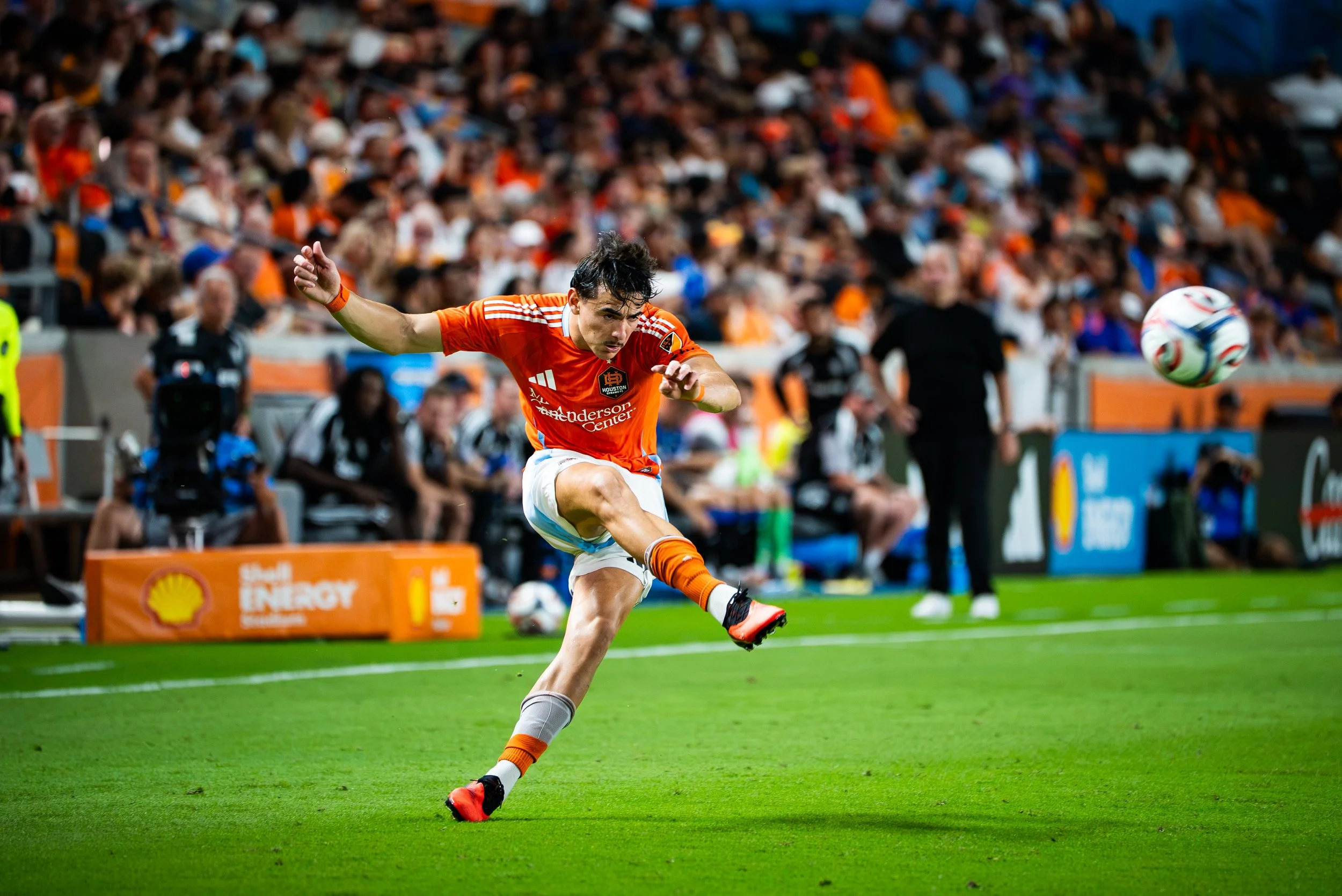 A male soccer player in an orange jersey and white shorts kicking a soccer ball on a green field during a game, with a crowd of spectators in the background.
