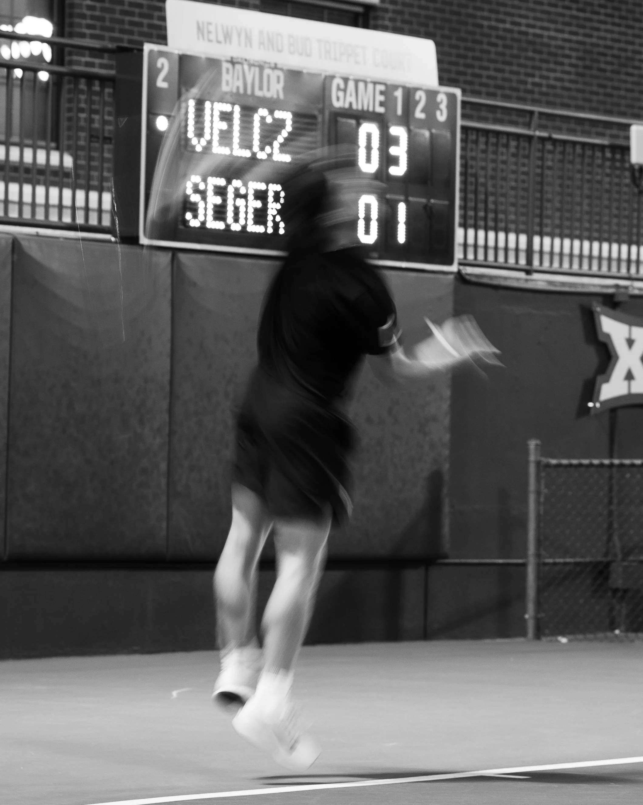 A blurred black and white photo of a person running on a track, with a scoreboard in the background displaying 'Baylor' and scores for game 1, 2, and 3.