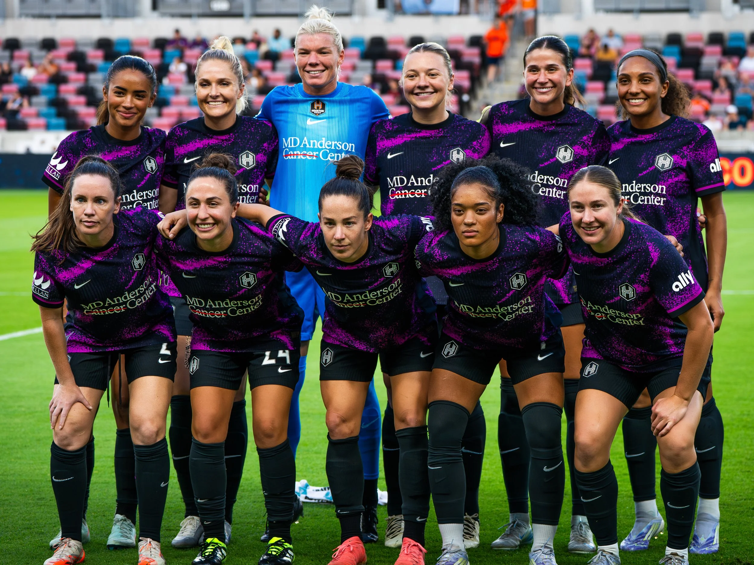 A women's soccer team in purple and black jerseys is posing for a team photo on a soccer field, with some players crouching and others standing. The player in the middle at the back is wearing a blue goalkeeper uniform. The background shows stadium s