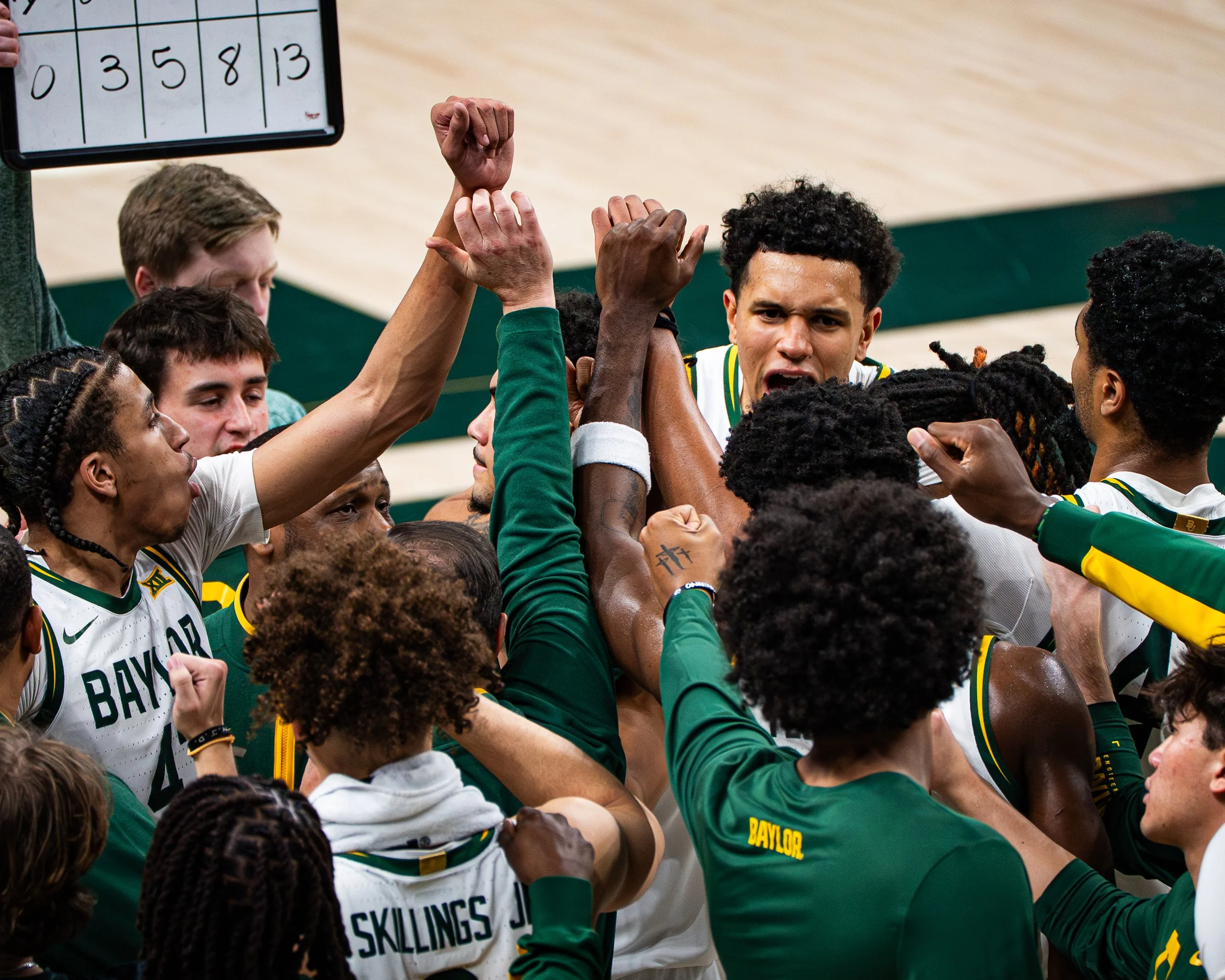 A group of basketball players in a huddle, raising their hands together, with a scoreboard visible in the background.